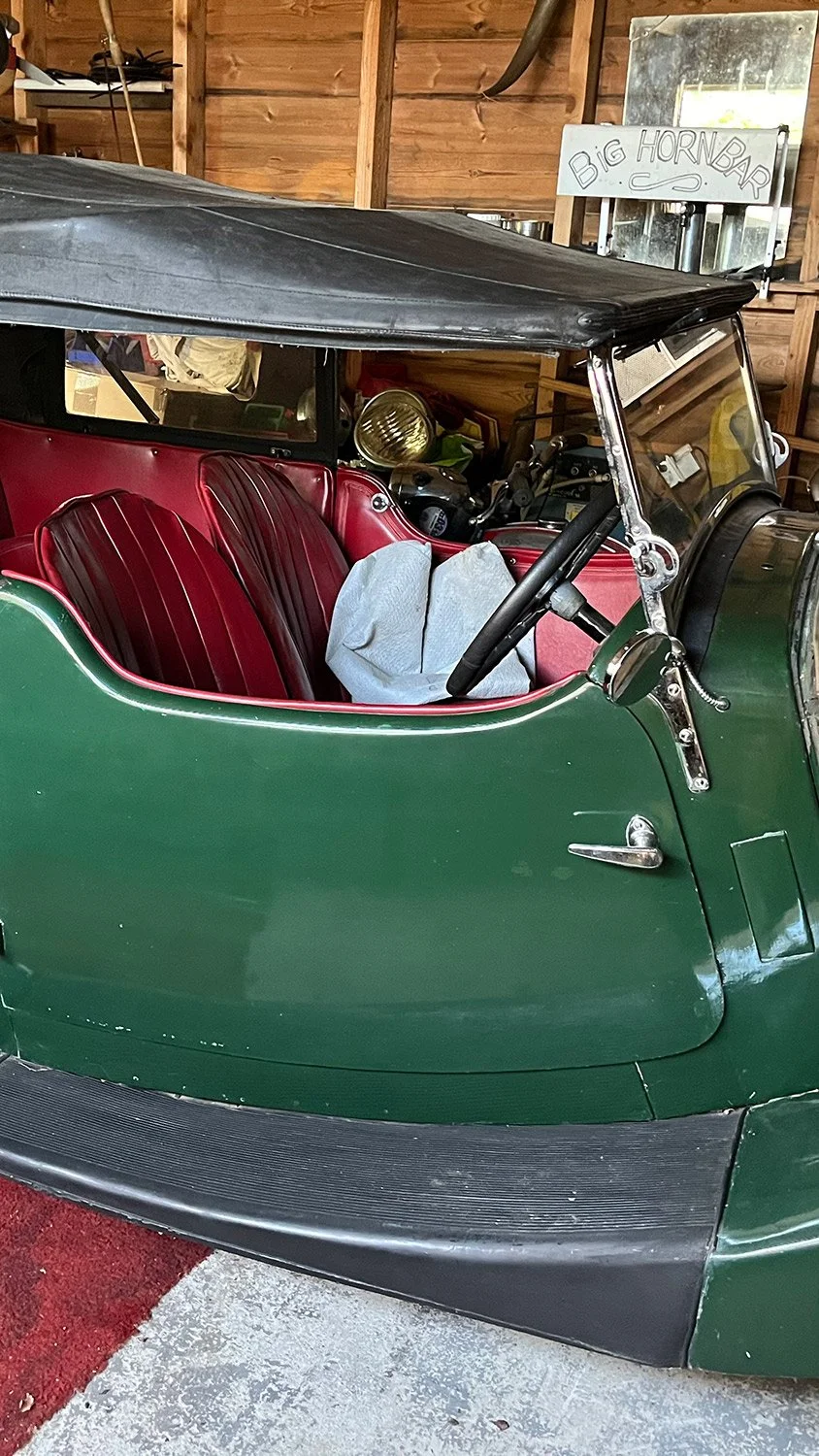 A vintage green car with red leather seats, parked inside a wooden garage with a sign that reads 'Big Horn Bar' in the background.