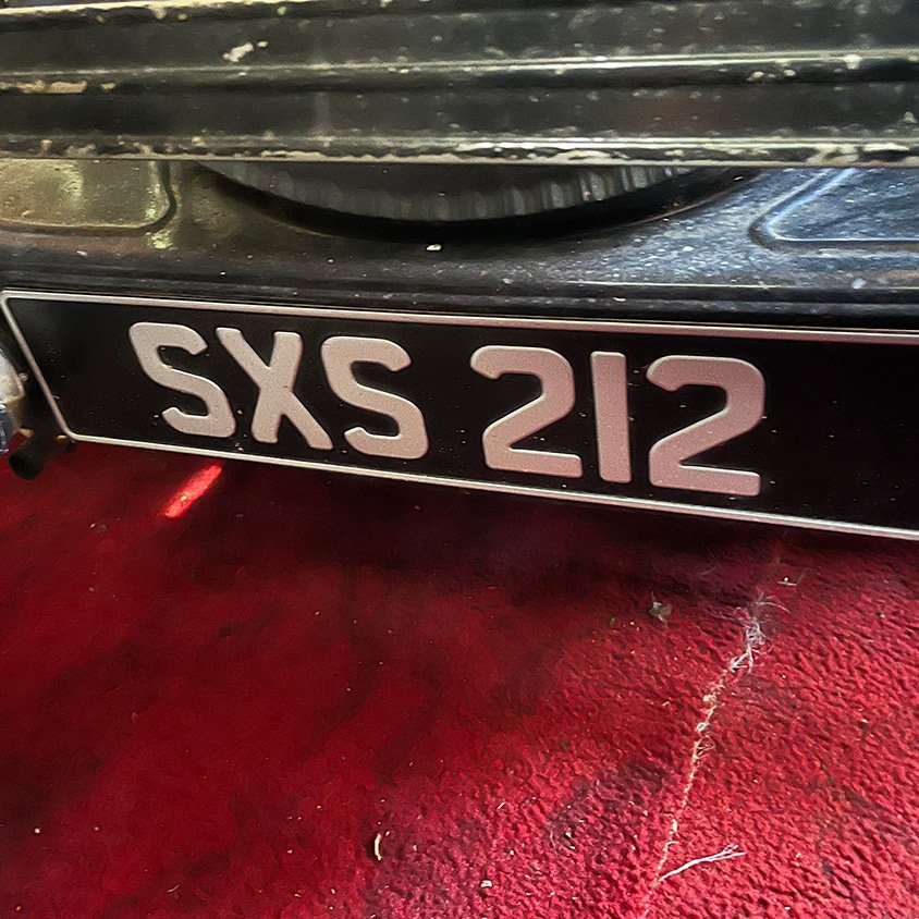 Close-up of a vintage black and white license plate reading 'SXS 212' mounted on a red surface.