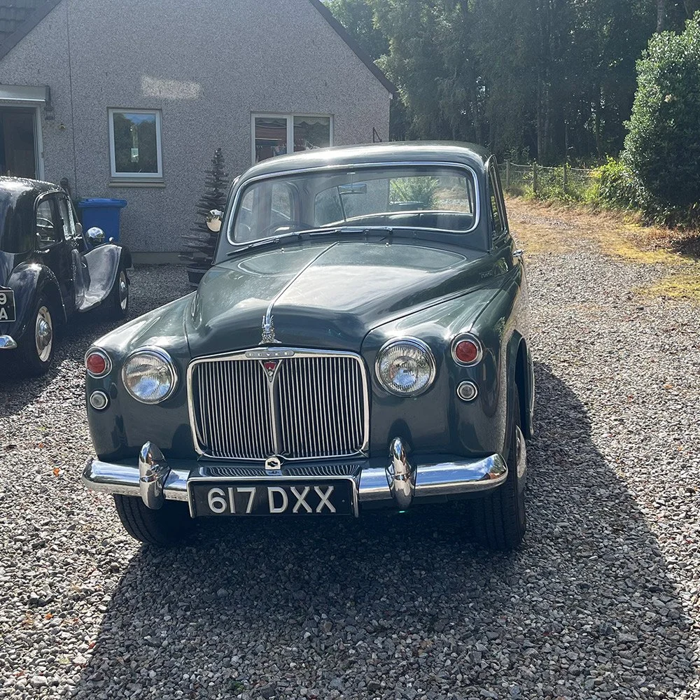 A vintage gray car parked on a gravel driveway in front of a house with trees in the background.