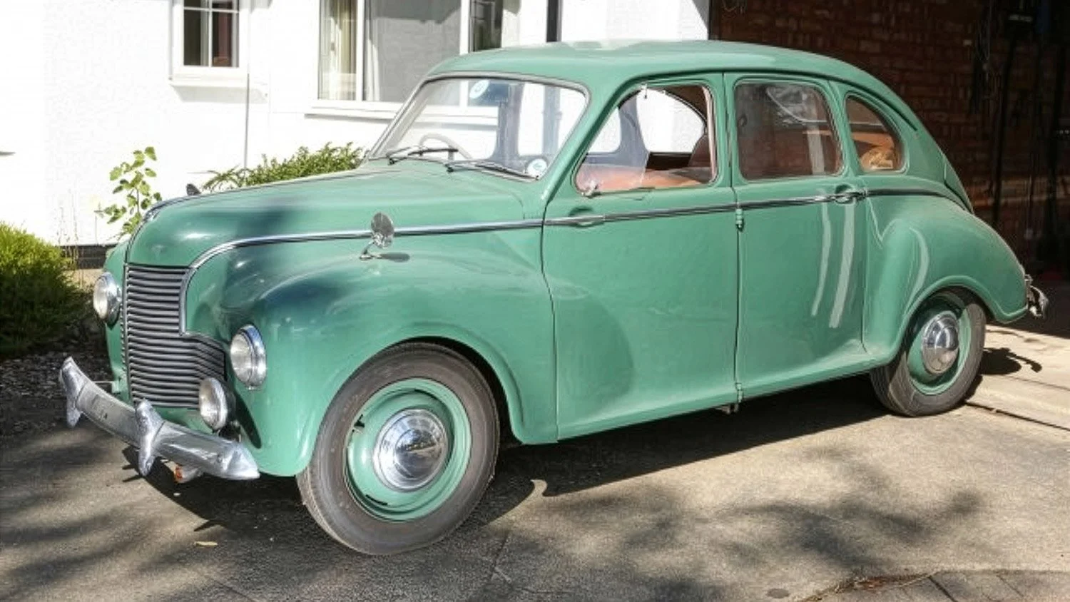 A vintage green car parked on a driveway near a white house and a brick wall.