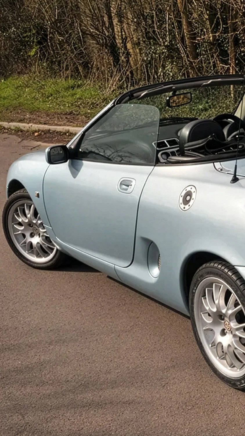 A silver convertible car parked on a paved road near a grassy area with trees in the background.