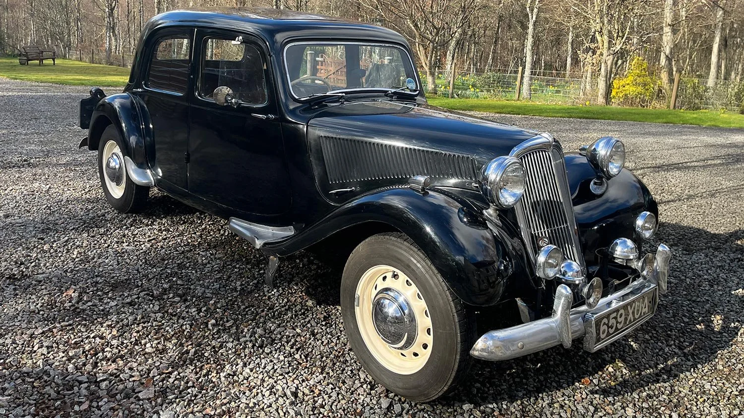 A vintage black car parked on a gravel surface with trees and a grassy area in the background.