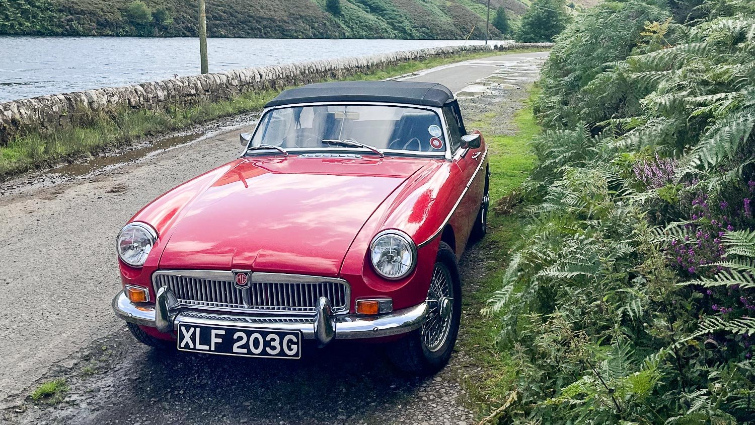 Red vintage MG convertible car parked on a gravel roadside near a body of water, with greenery and flowering plants nearby.