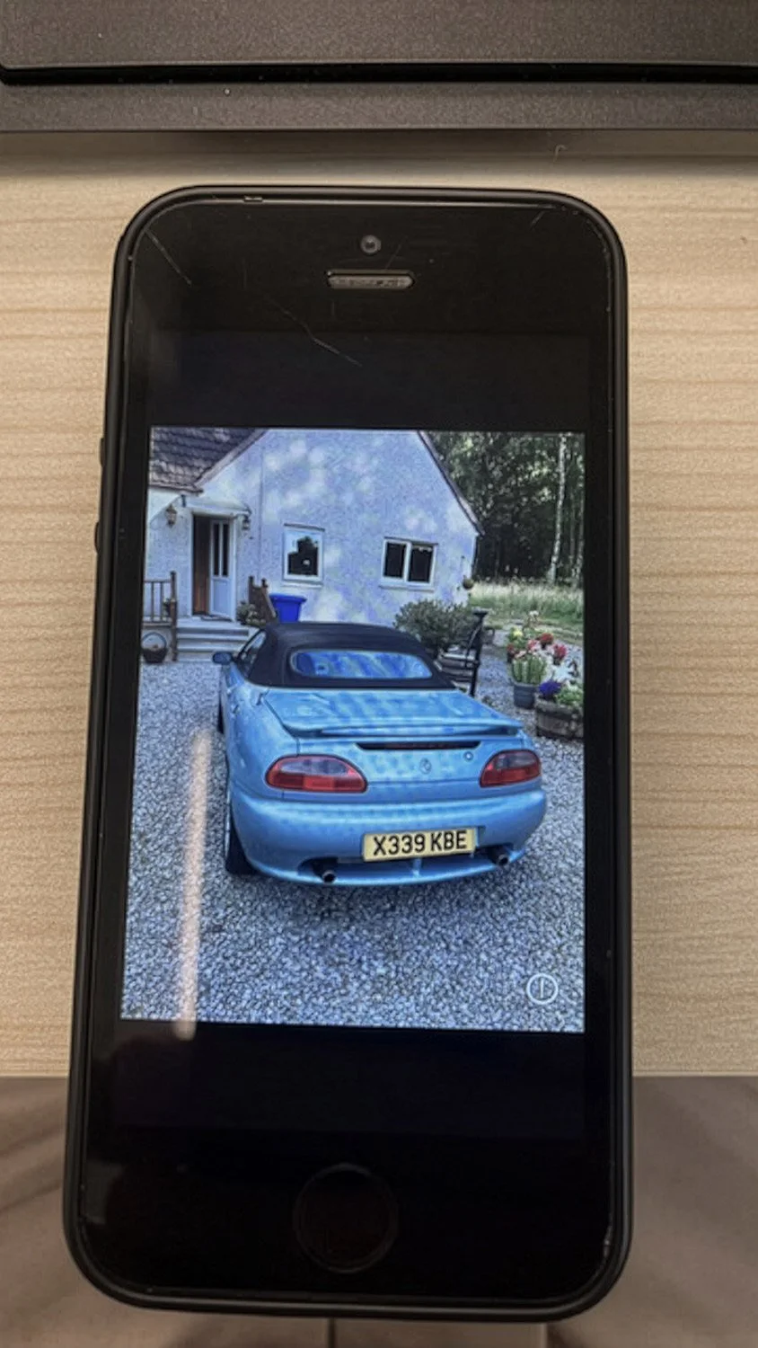 A smartphone showing a photo of a blue convertible car parked in front of a house with white walls, two windows, and a small porch, surrounded by a gravel driveway and some outdoor plants.