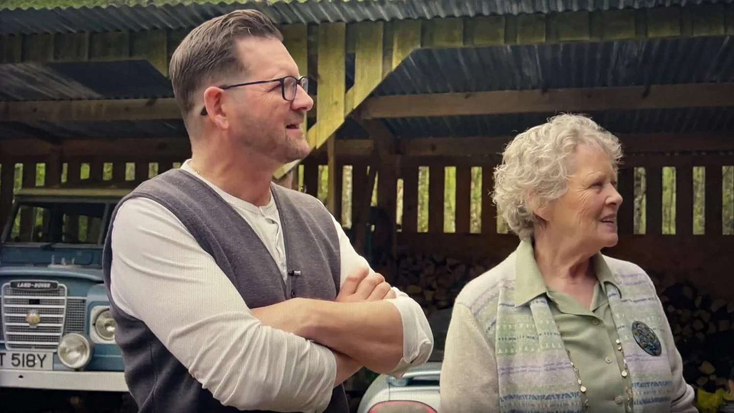 John and his mother standing outdoors in front of a wooden structure, smiling and talking. Mrs Mackenzie wearing a perched vest and a broach, and John wearing a beige shirt and grey waistcoat.