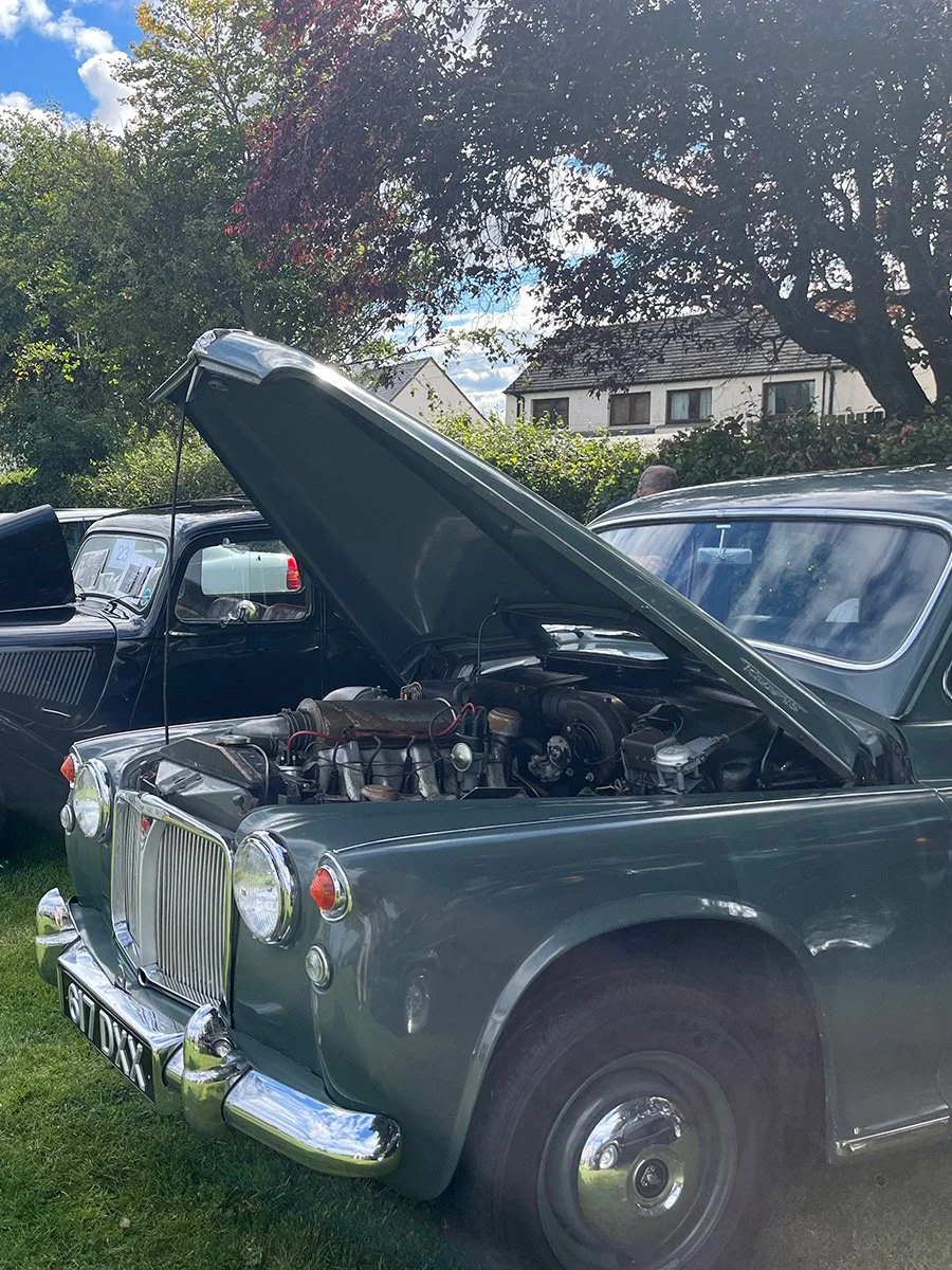 A vintage gray car with its hood open at a classic car show, parked on grass with other cars and trees in the background.