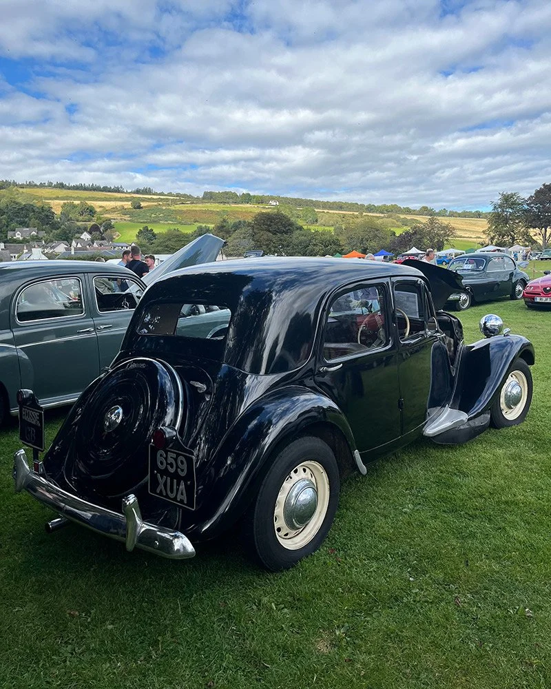 A vintage black car parked on a grassy field at a classic car show, with other classic cars and people in the background, under a partly cloudy sky.