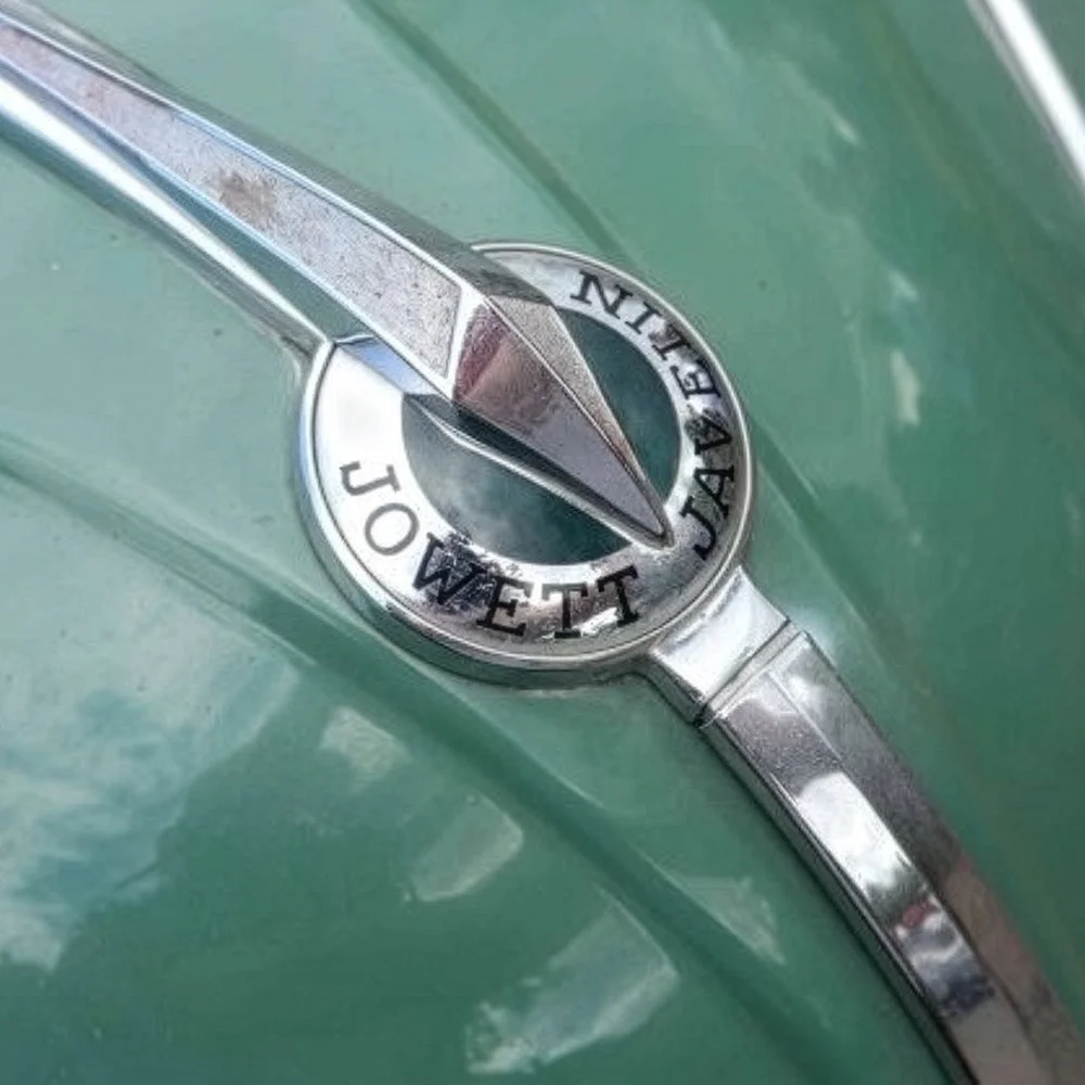 Close-up of a vintage green car hood ornament with a circular badge that reads "JOWELL" surrounding a stylized chrome design.