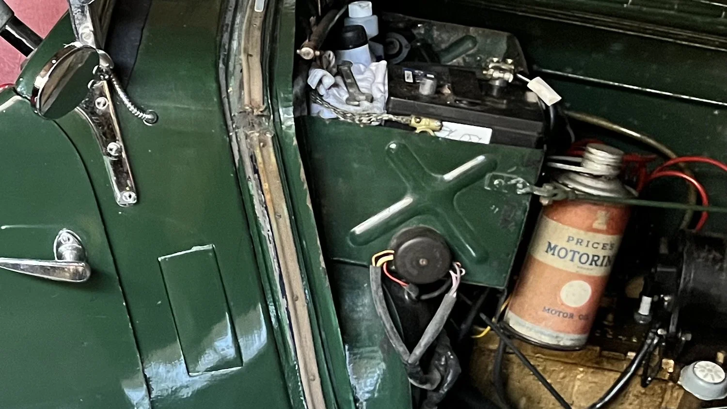 Close-up of a vintage car engine compartment with a green exterior, showing parts like a coil, wires, and an old motor oil canister.