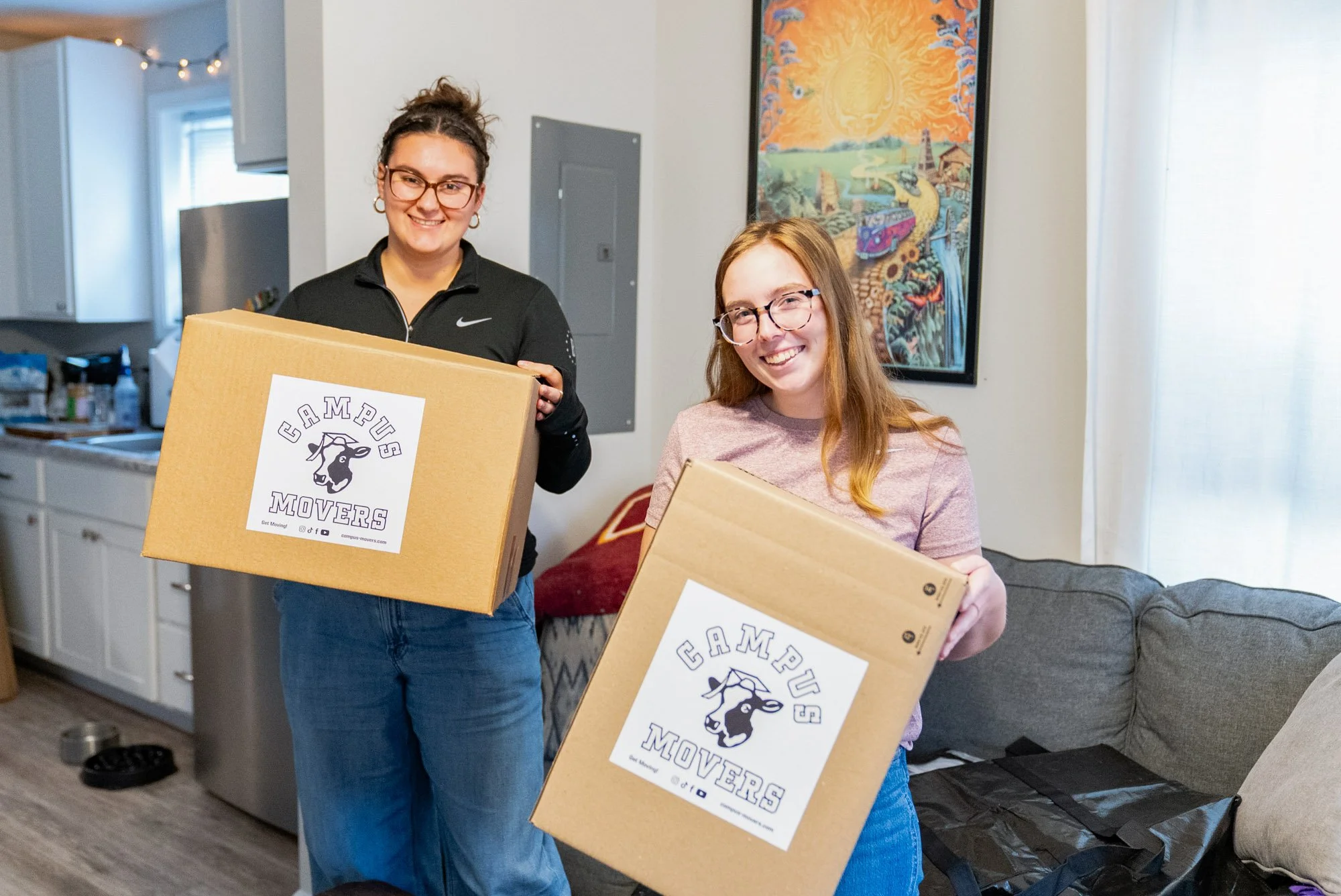Two women smiling and holding cardboard boxes with 'Campus Movers' labels, standing in a living room.