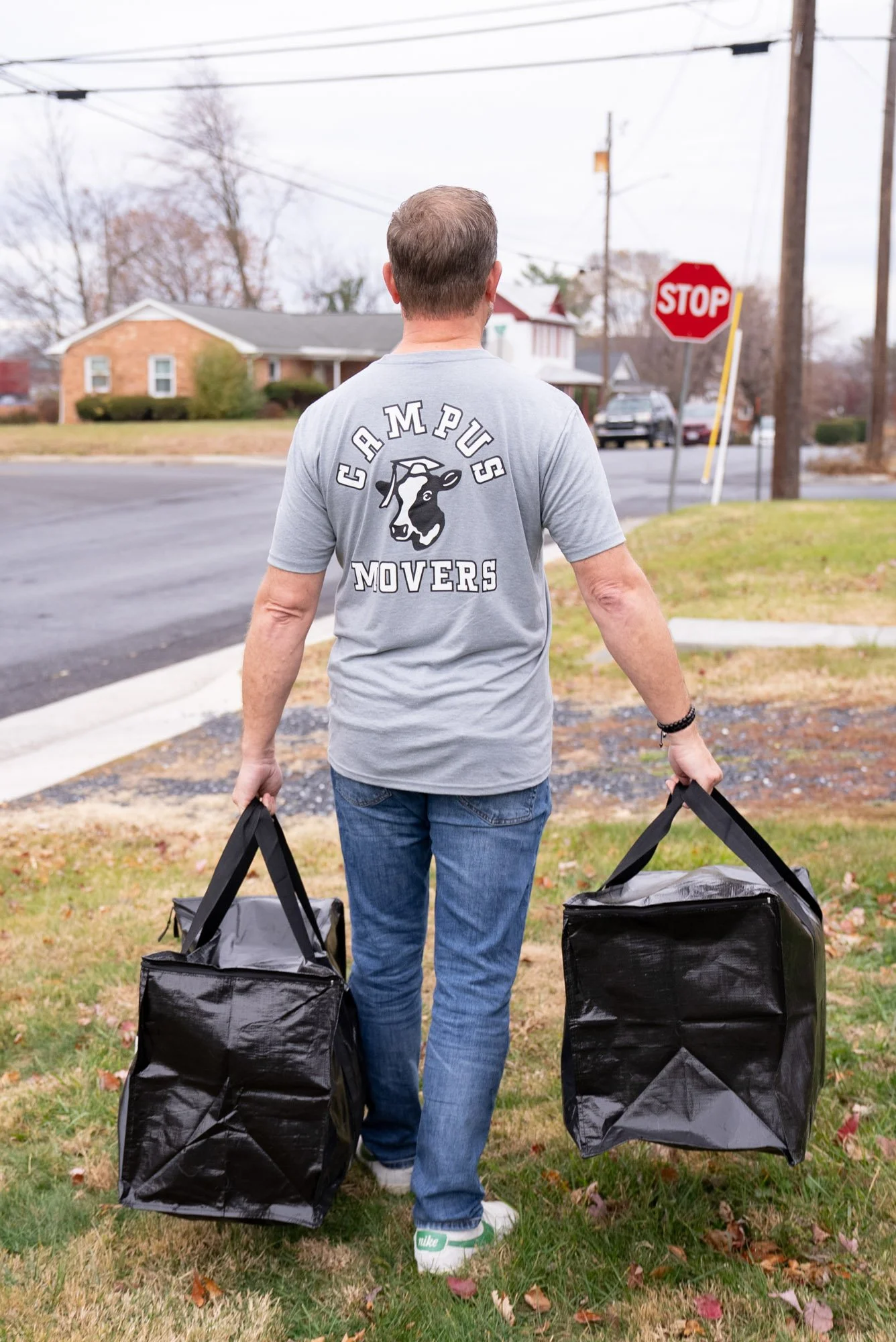 A man carrying two black insulated bags, walking away from the camera near a residential street with an overcast sky.