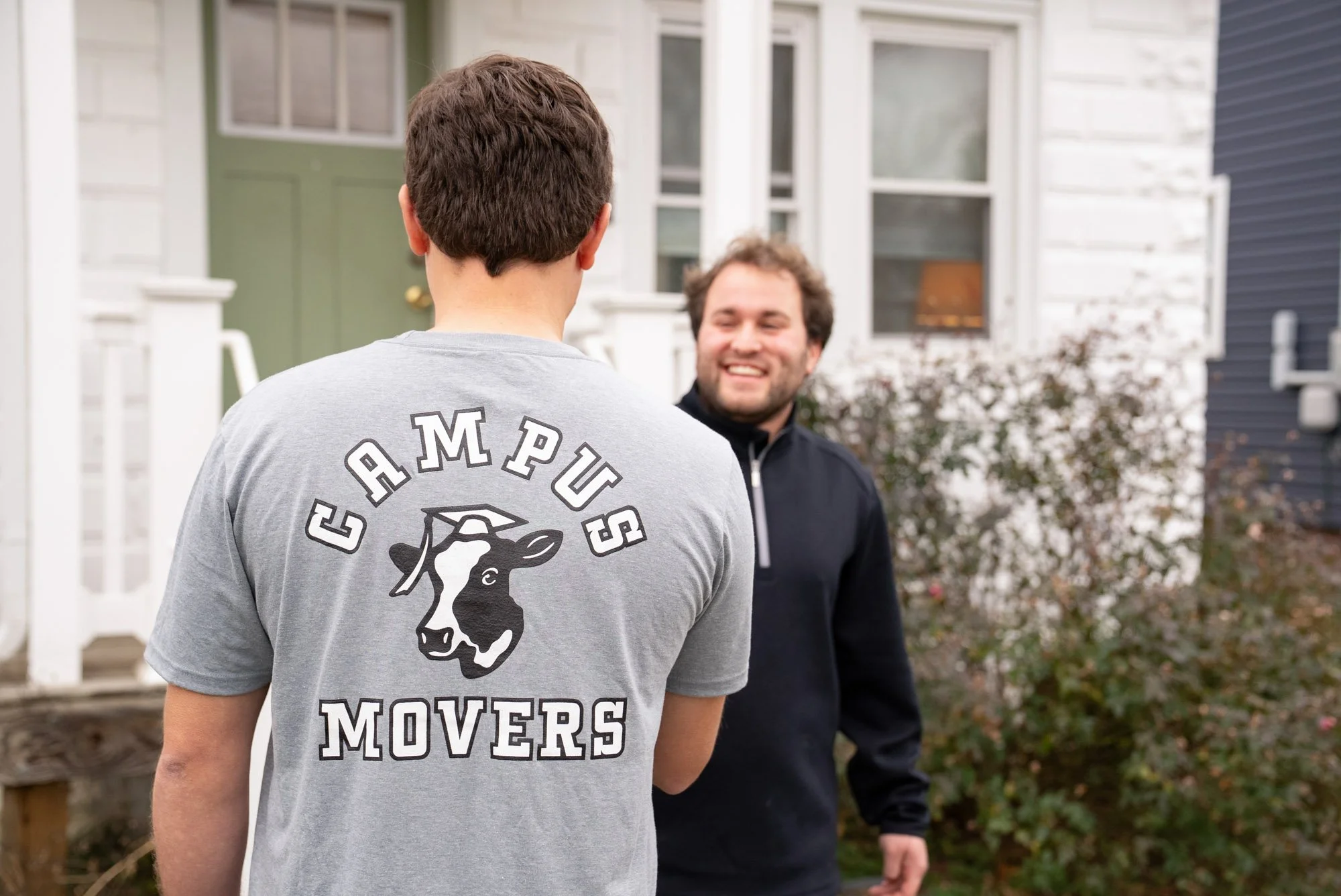 Two men conversing outside of a house. One person is facing away from the camera, wearing a gray t-shirt with 'CAMPUS MOVERS' and a cow logo. The other man is smiling, wearing a black zip-up jacket, and facing the person with the t-shirt.