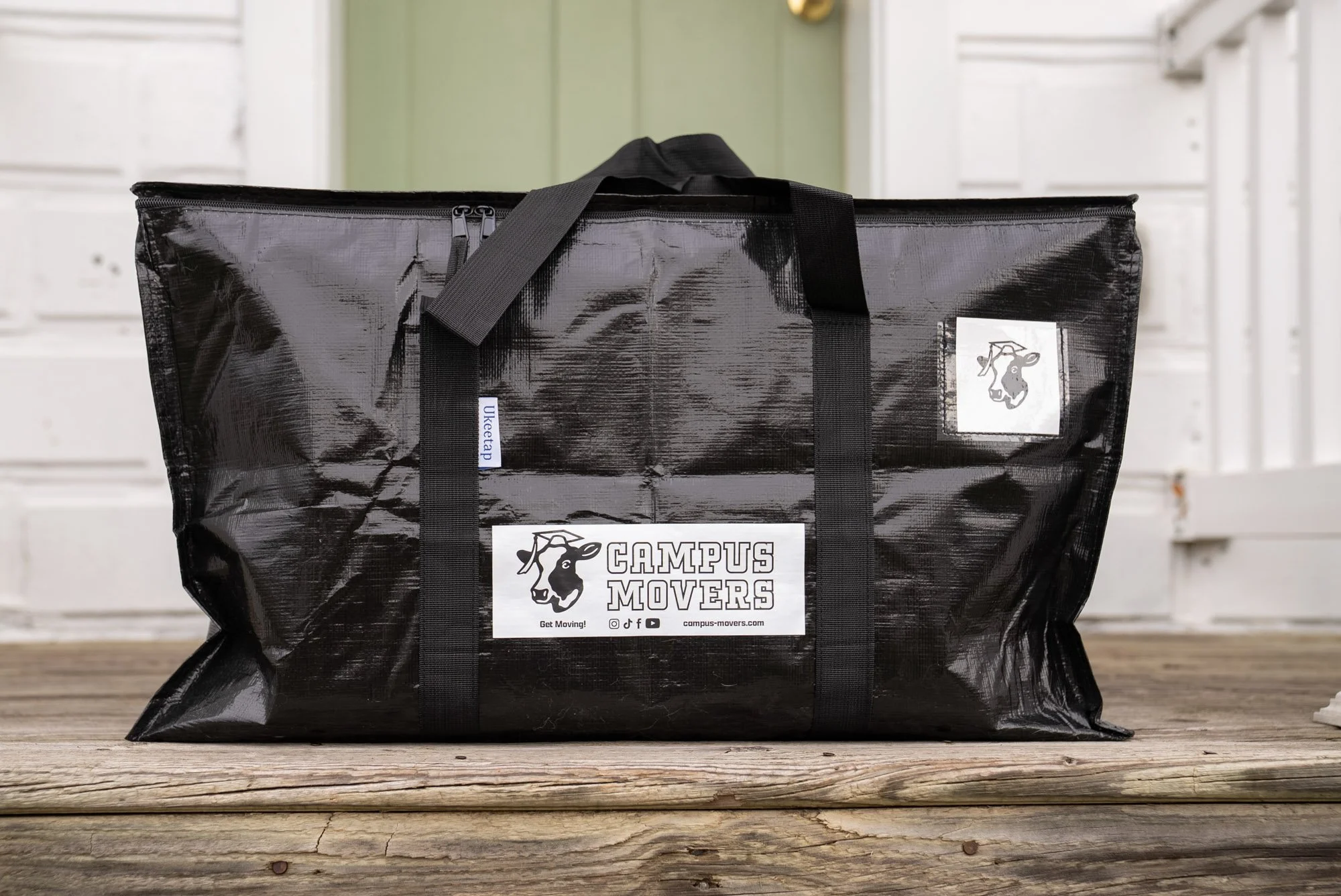 Black reusable moving bag with handles, labeled 'Campus Movers' on a wooden surface in front of white and green background.