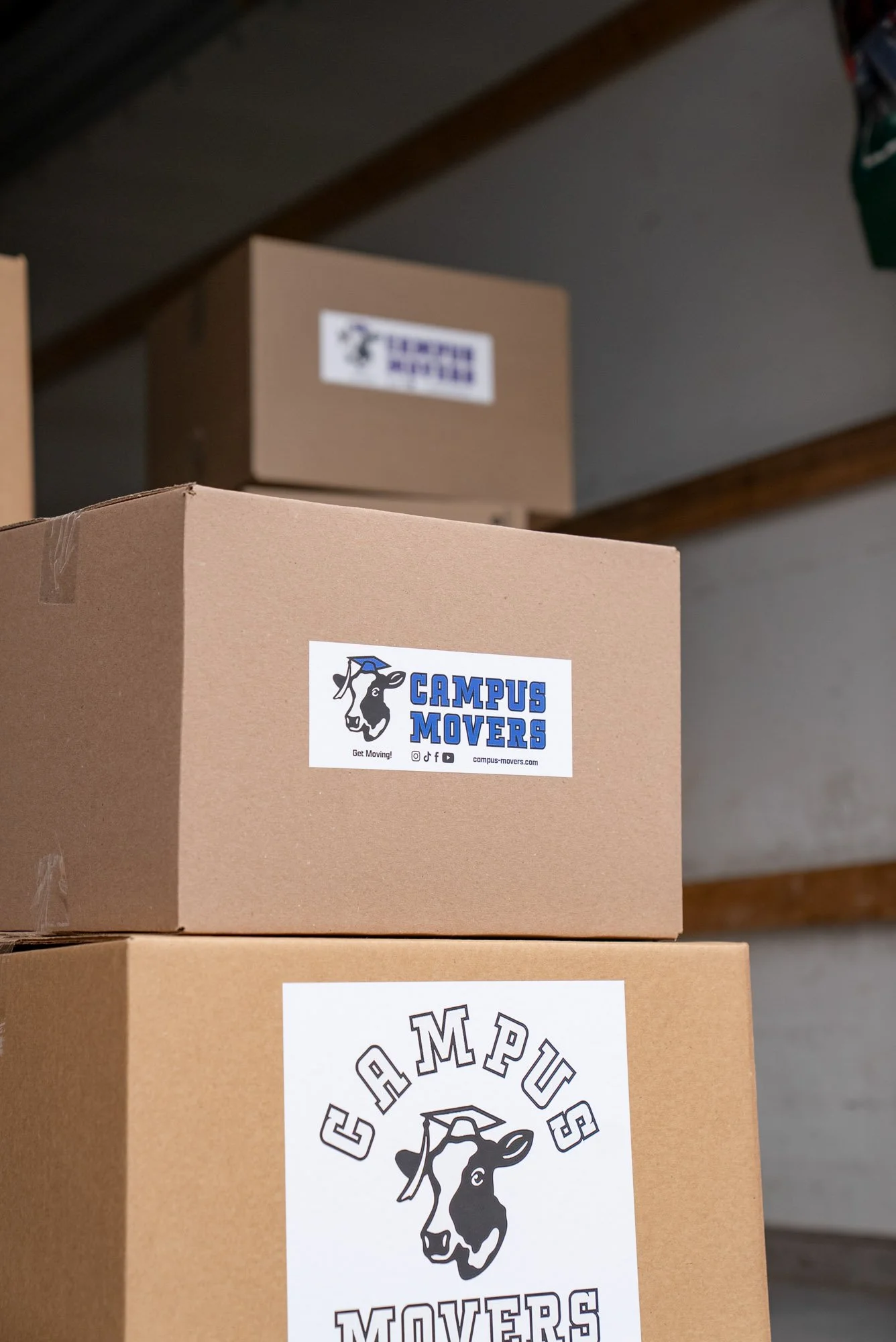 Stack of cardboard boxes with Campus Movers labels, one with a logo of a cow wearing a graduation cap, placed on a shelf in a storage room.