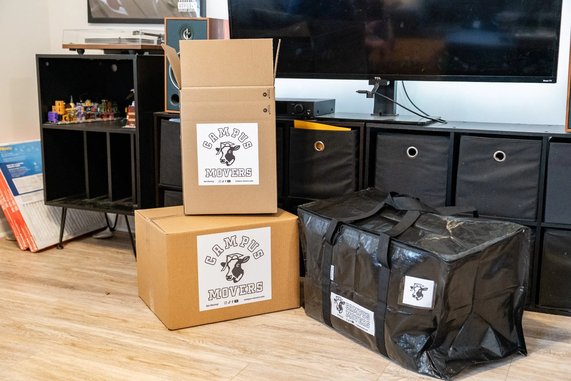 Boxes and a large black bag labeled 'Campus Movers' in a room with wooden flooring, black shelving units, and a flat-screen TV.