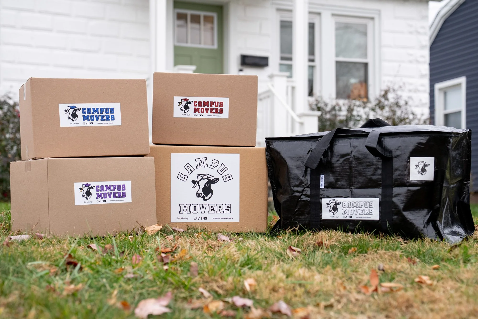 Boxes and a black bag with Campus Movers logos on the front yard of a house.