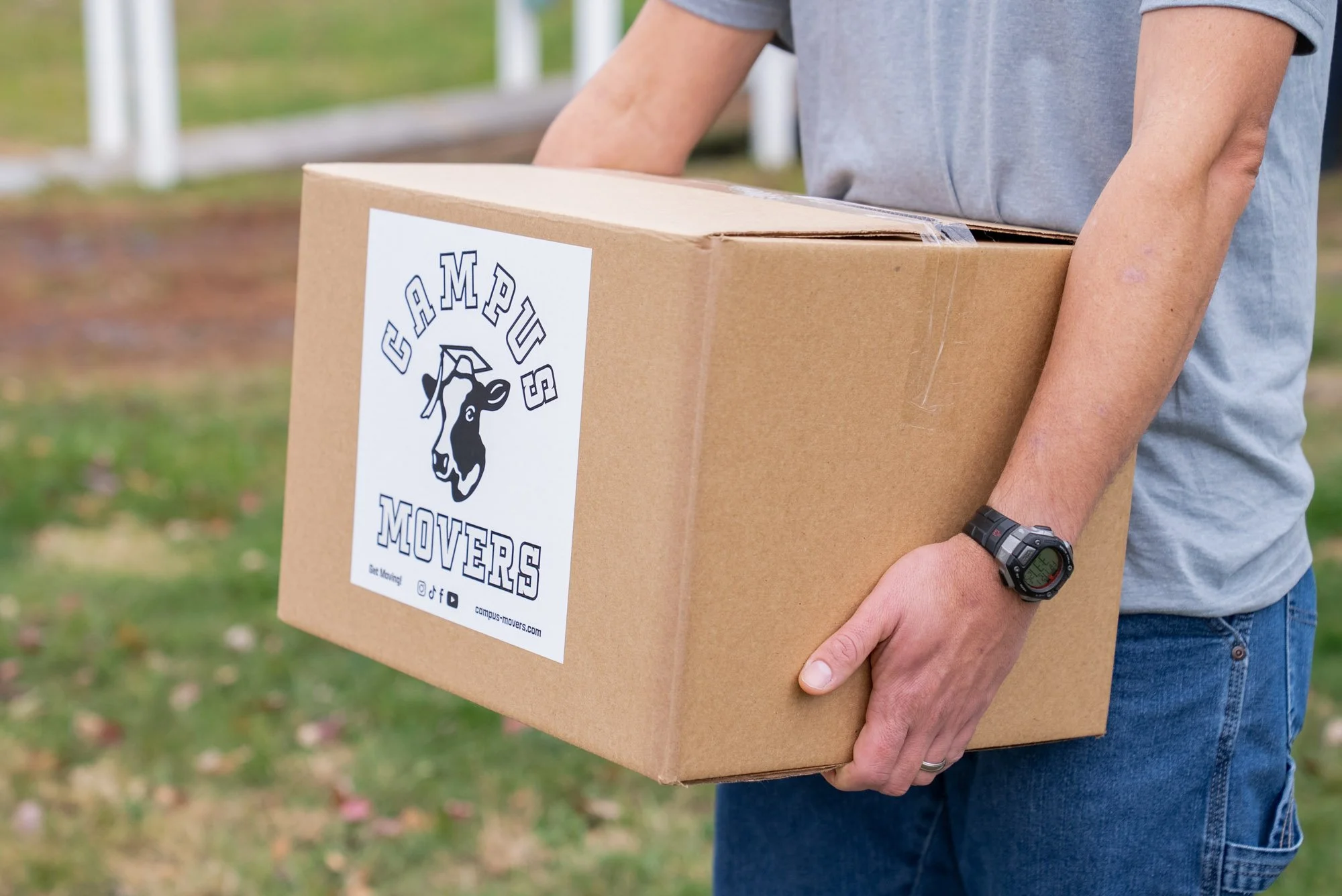 Person holding a cardboard box with a 'Campus Movers' logo on it, outdoors on grass with some fallen leaves.