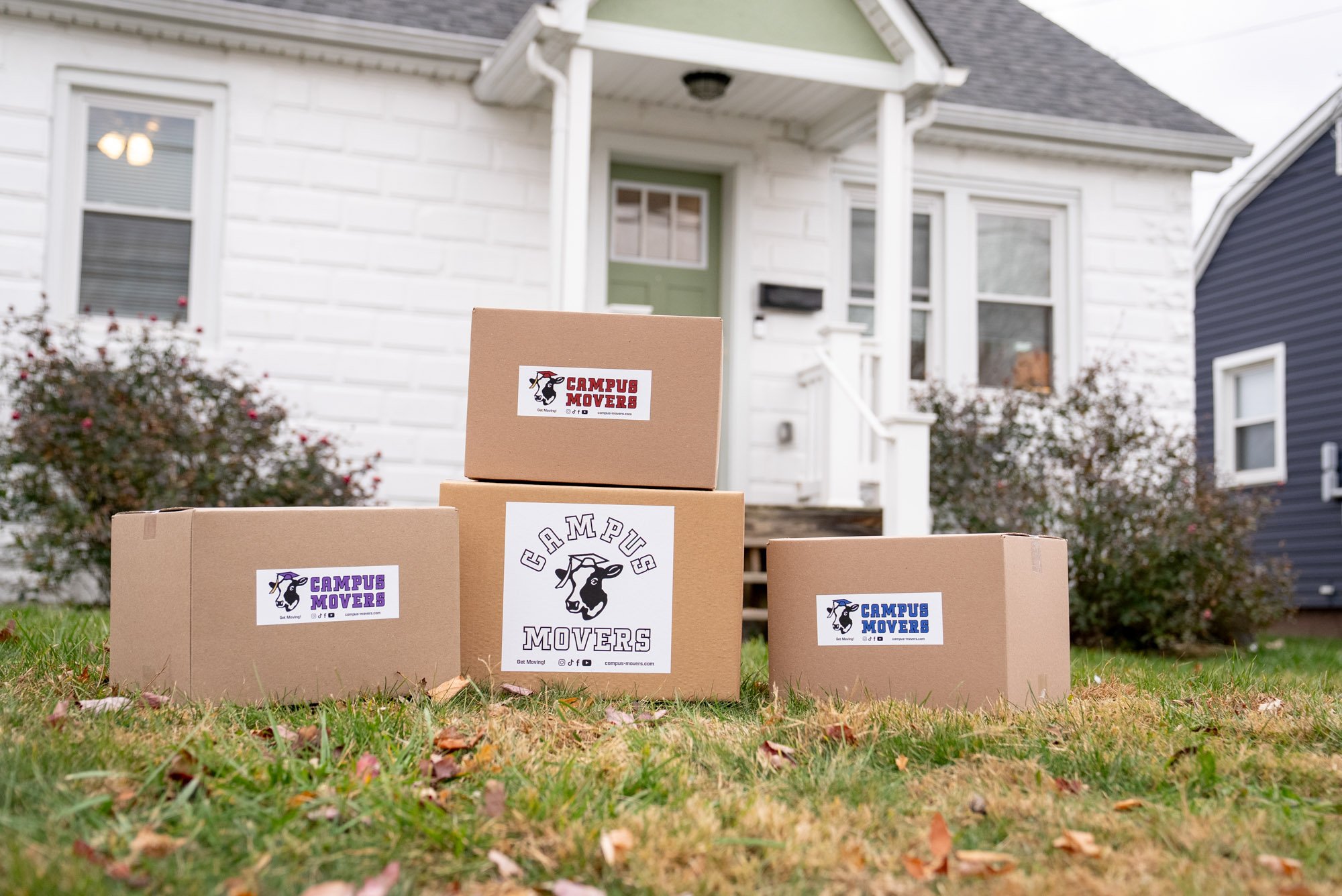 Four cardboard boxes with 'Campus Movers' labels are placed on a lawn in front of a white house with a green door. Two boxes are labeled purple, one red, and one blue.