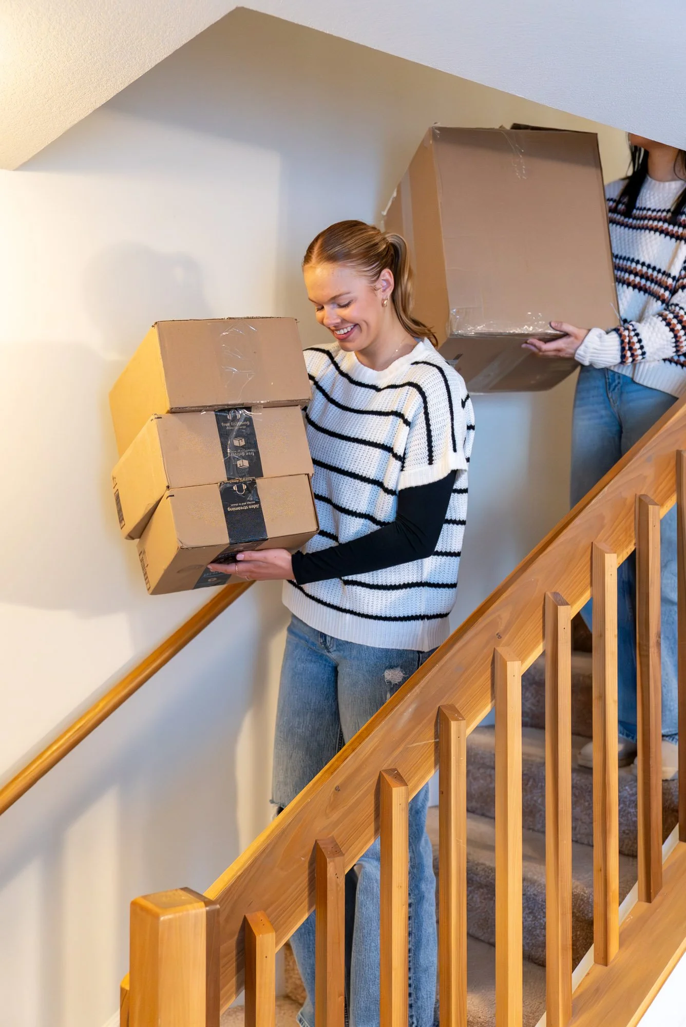Two women carrying Amazon packages down a wooden staircase, one woman holding three boxes stacked in her hands and the other woman holding a larger box.
