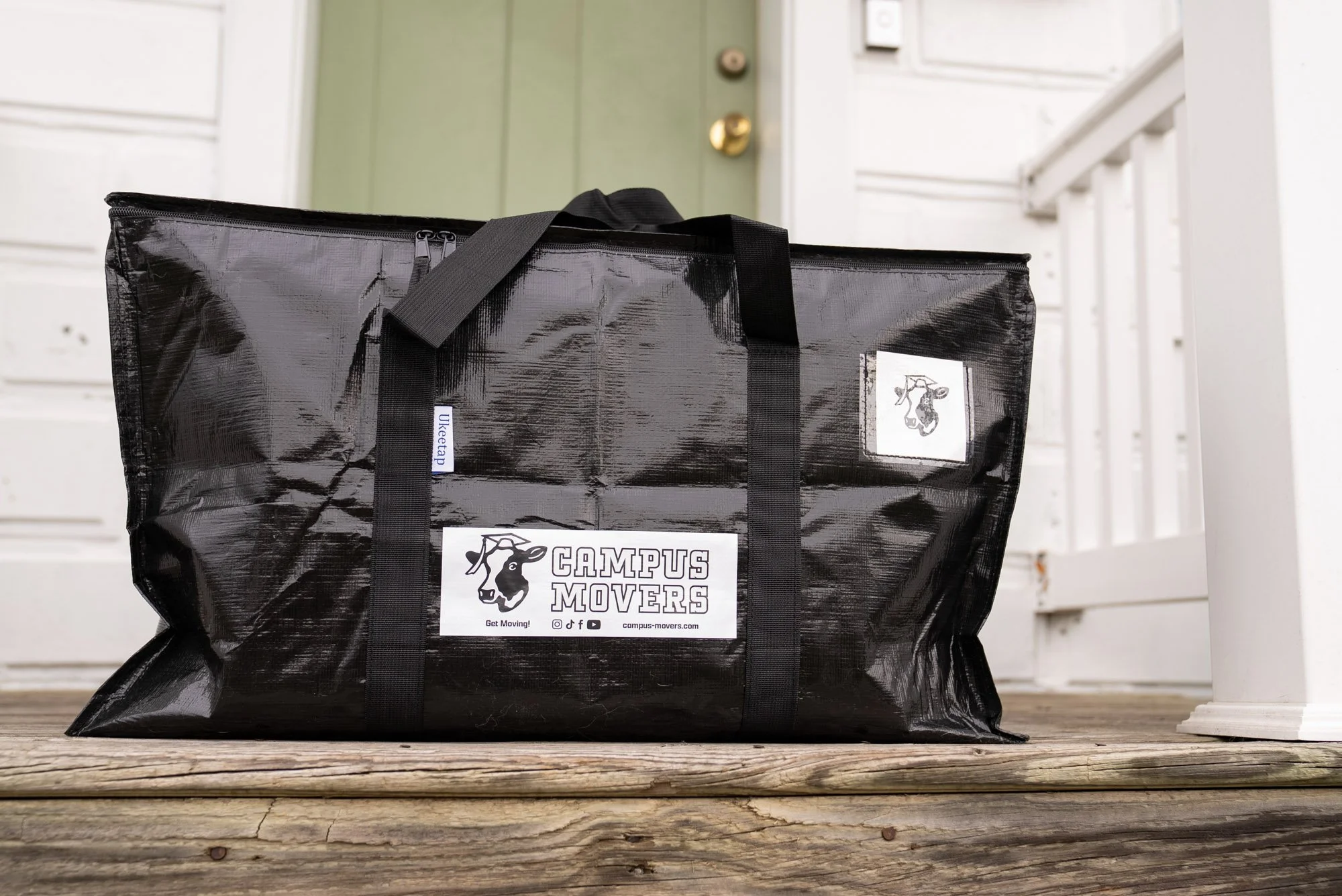 A large black tote bag with handles sitting on a wooden porch floor. The bag has a white logo sticker that reads 'CAMPUS MOVERS' with a cartoon cow, social media icons, and a website address. A small white label on the bag reads 'UncrateP'. In the ba