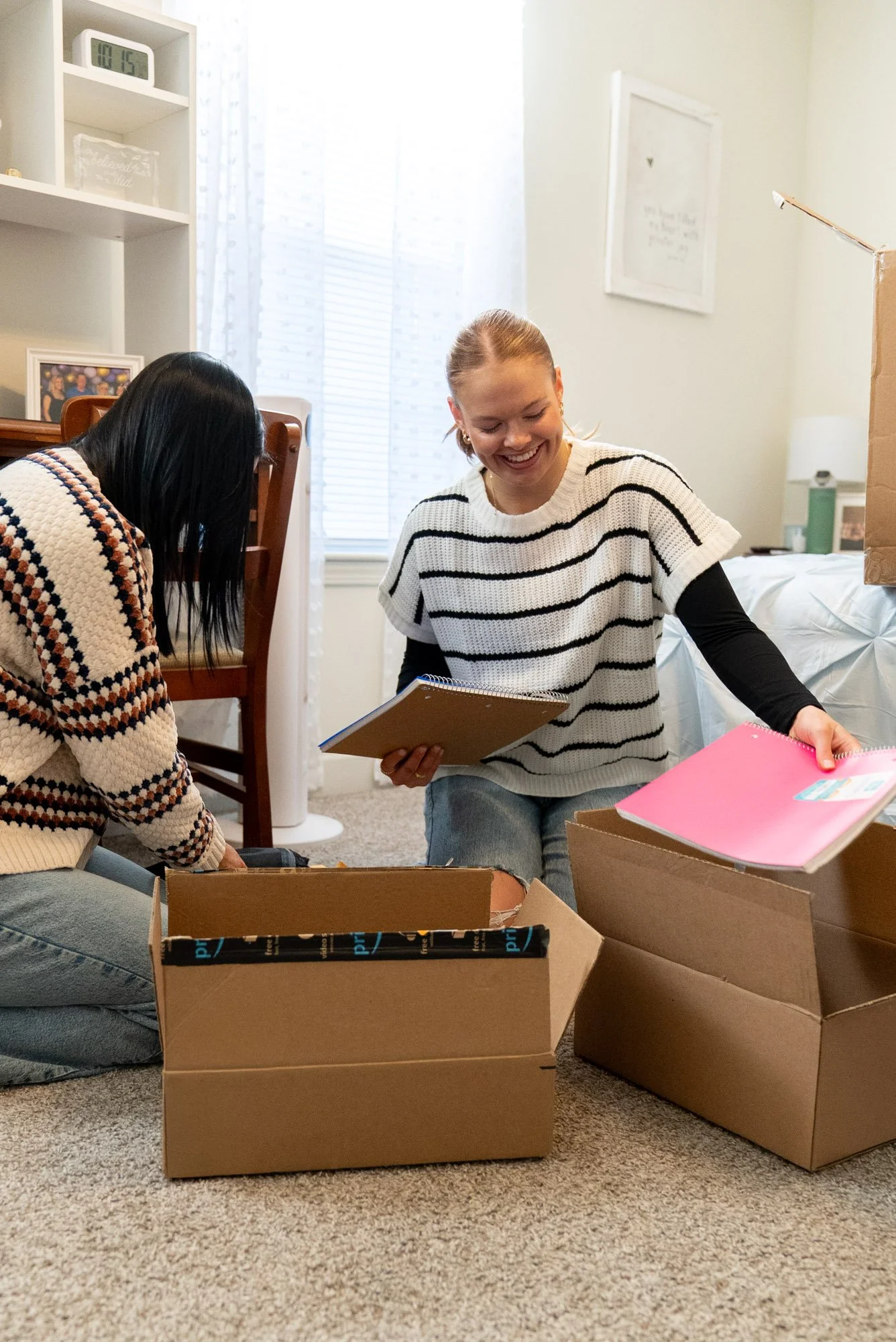 Two women unpacking moving boxes inside a home, smiling and holding notebooks, with a window and a framed picture on the wall in the background.