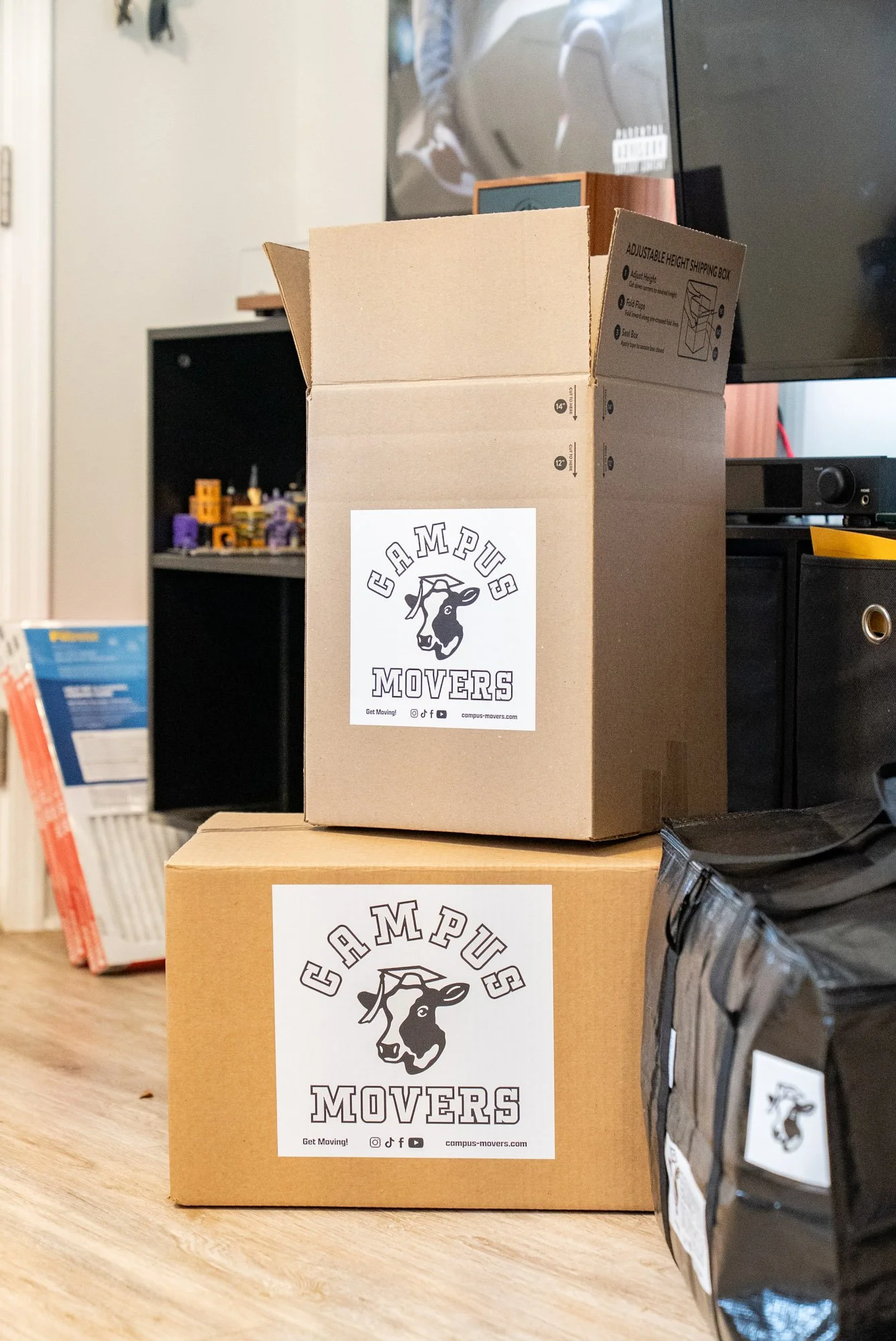 Two cardboard boxes with 'Campus Movers' stickers on a wooden floor, one on top of the other, near a black bag and a black shelf in a room
