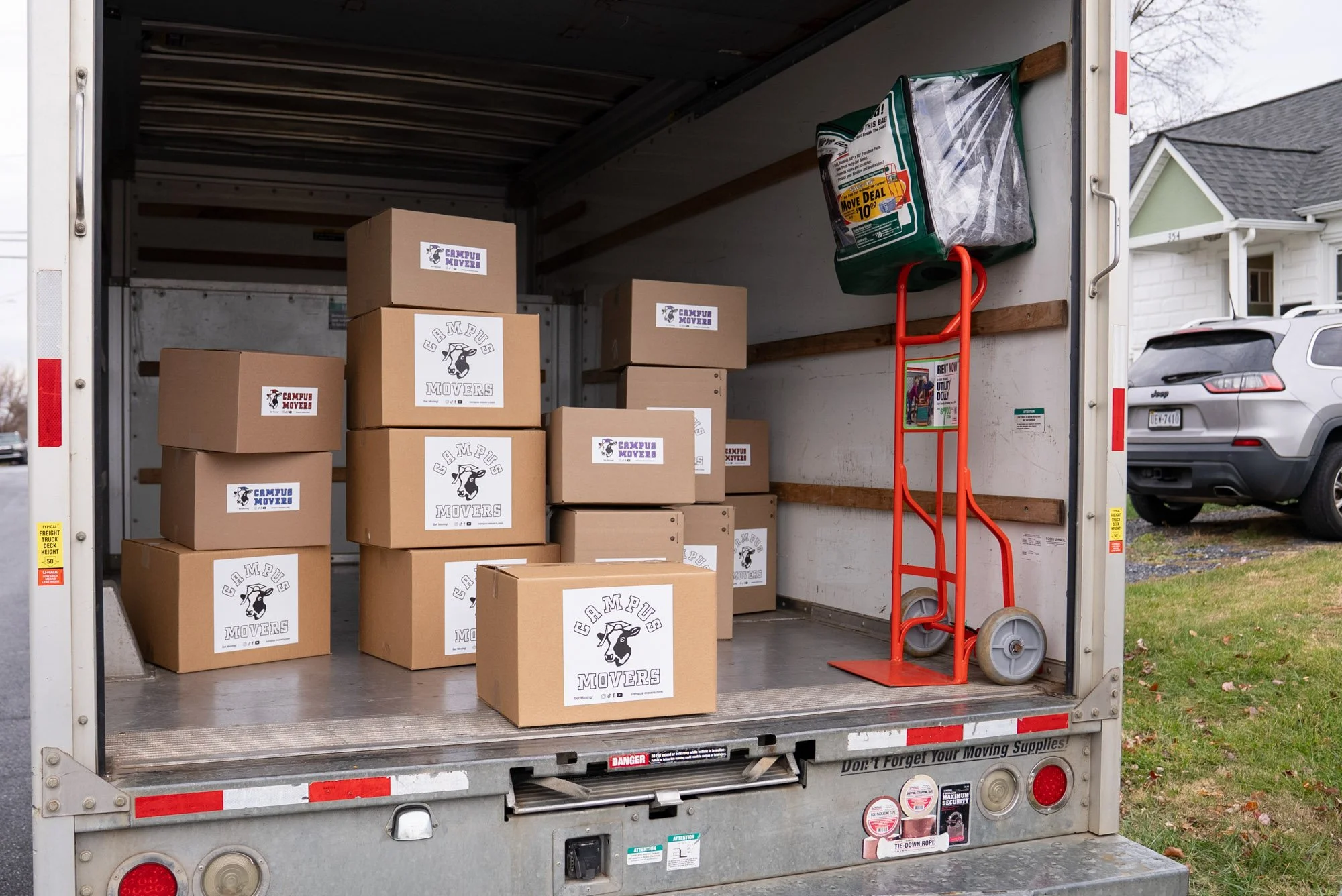 Open moving truck with cardboard boxes labeled 'Campus Movers' inside, a red hand truck leaning against the wall, and a green moving blanket pushed into the corner.