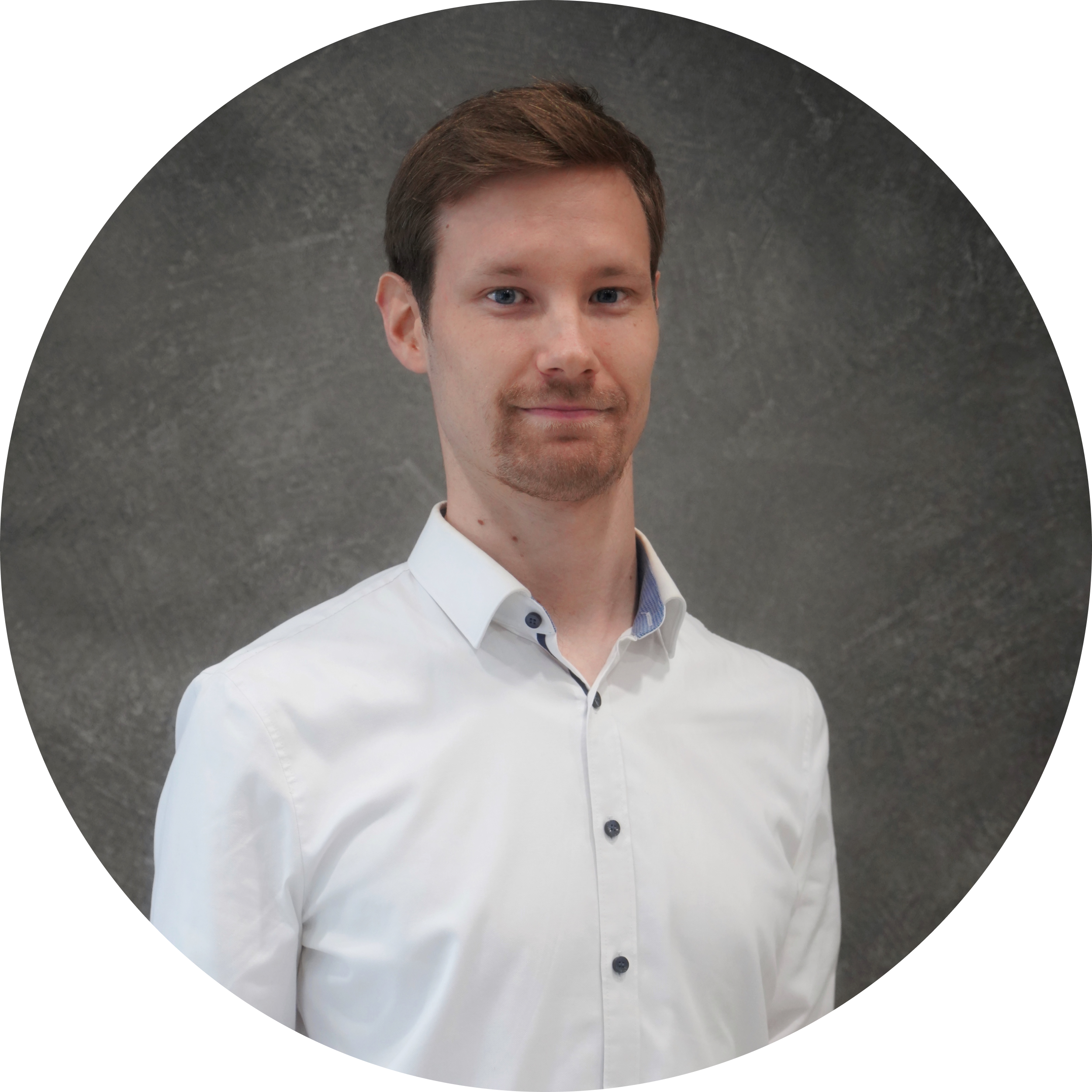 Portrait of a young man with light brown hair, blue eyes, and a trimmed beard, wearing a white button-up shirt, standing against a grey textured background.