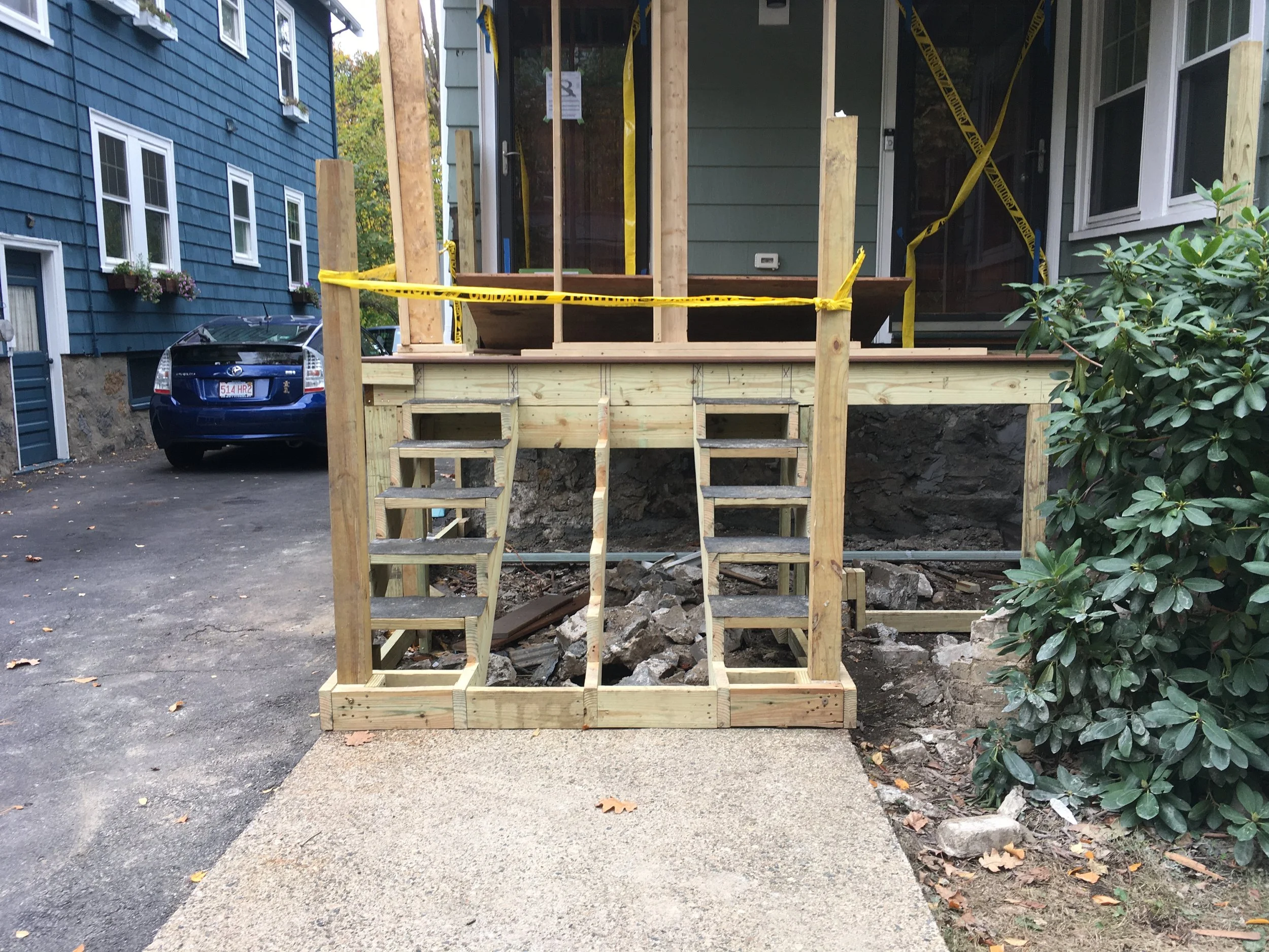 Front view of a house with a new wooden porch construction, stairs, and supporting posts; caution tape surrounds the area, and there is a driveway with a parked blue car on the left.