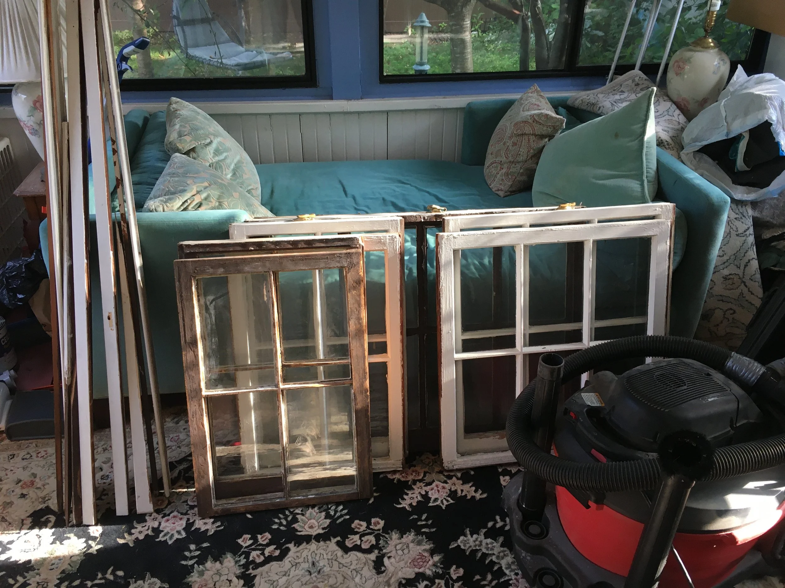 A living room with a turquoise sofa and pillows, multiple window panes leaning against the sofa, and a vacuum cleaner on a floral rug.