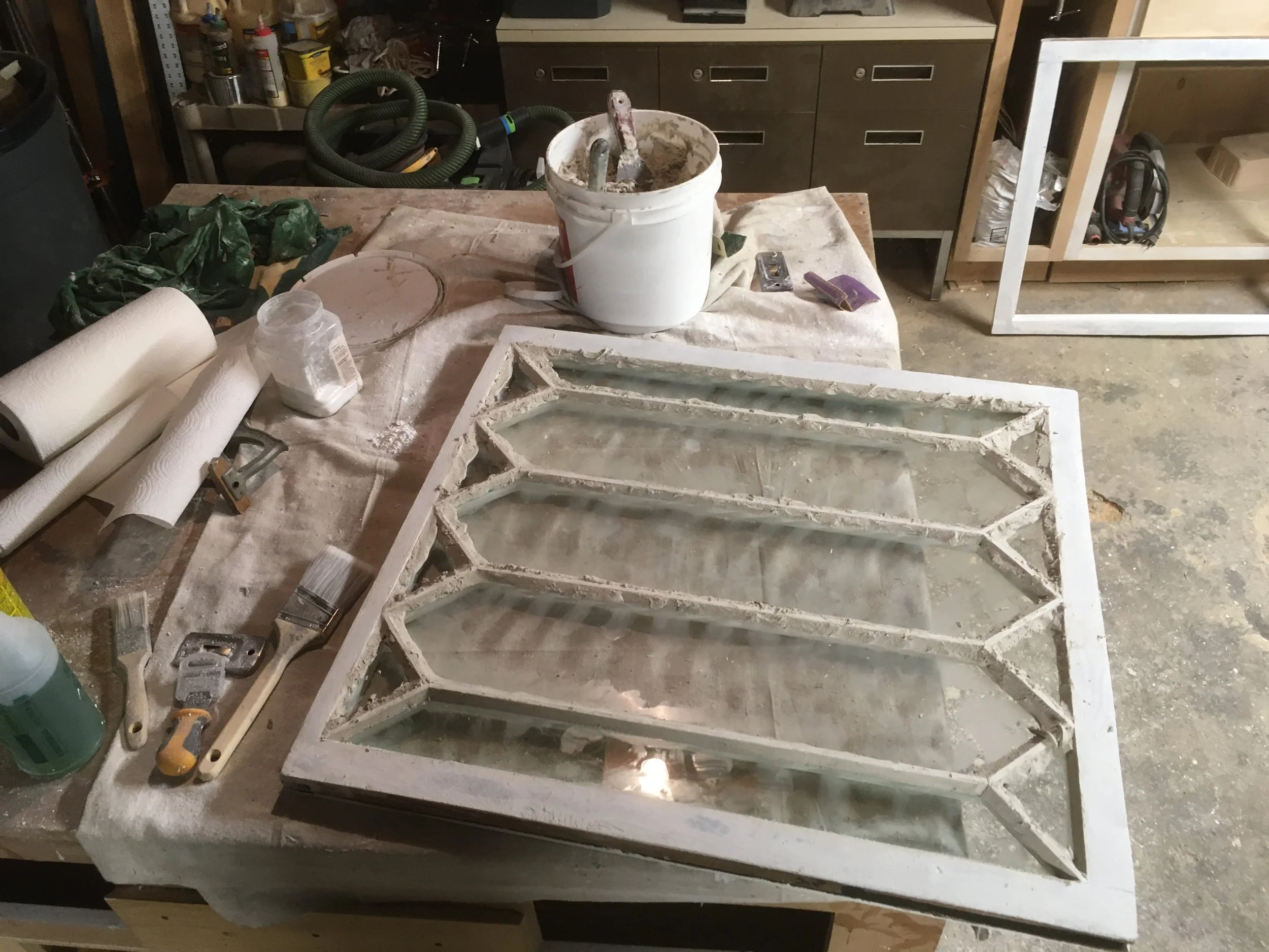 A workshop table with a large, unfinished stained glass window frame. The frame has several geometric sections, some with glass panes, and is surrounded by tools, paint brushes, and supplies like glue, paint, and rags.