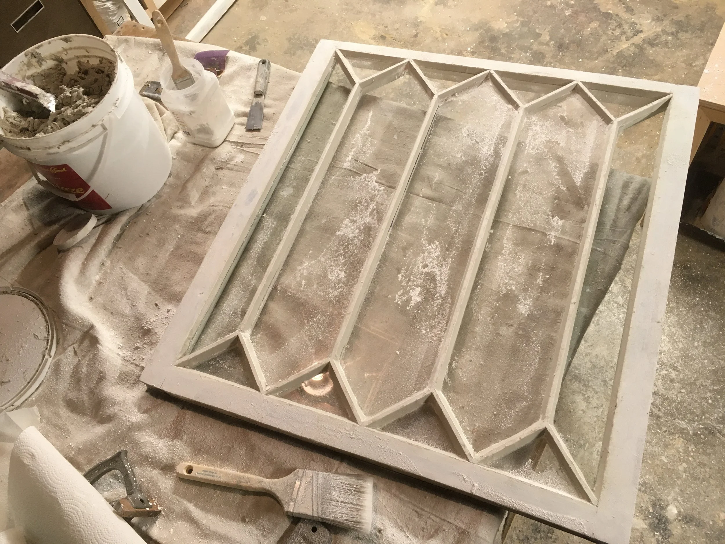 A wooden frame with geometric patterns is being crafted on a work table. The table is covered in dust, and there are various tools and supplies, including a bucket, a paintbrush, and a container of cement or plaster, indicating an ongoing woodworking