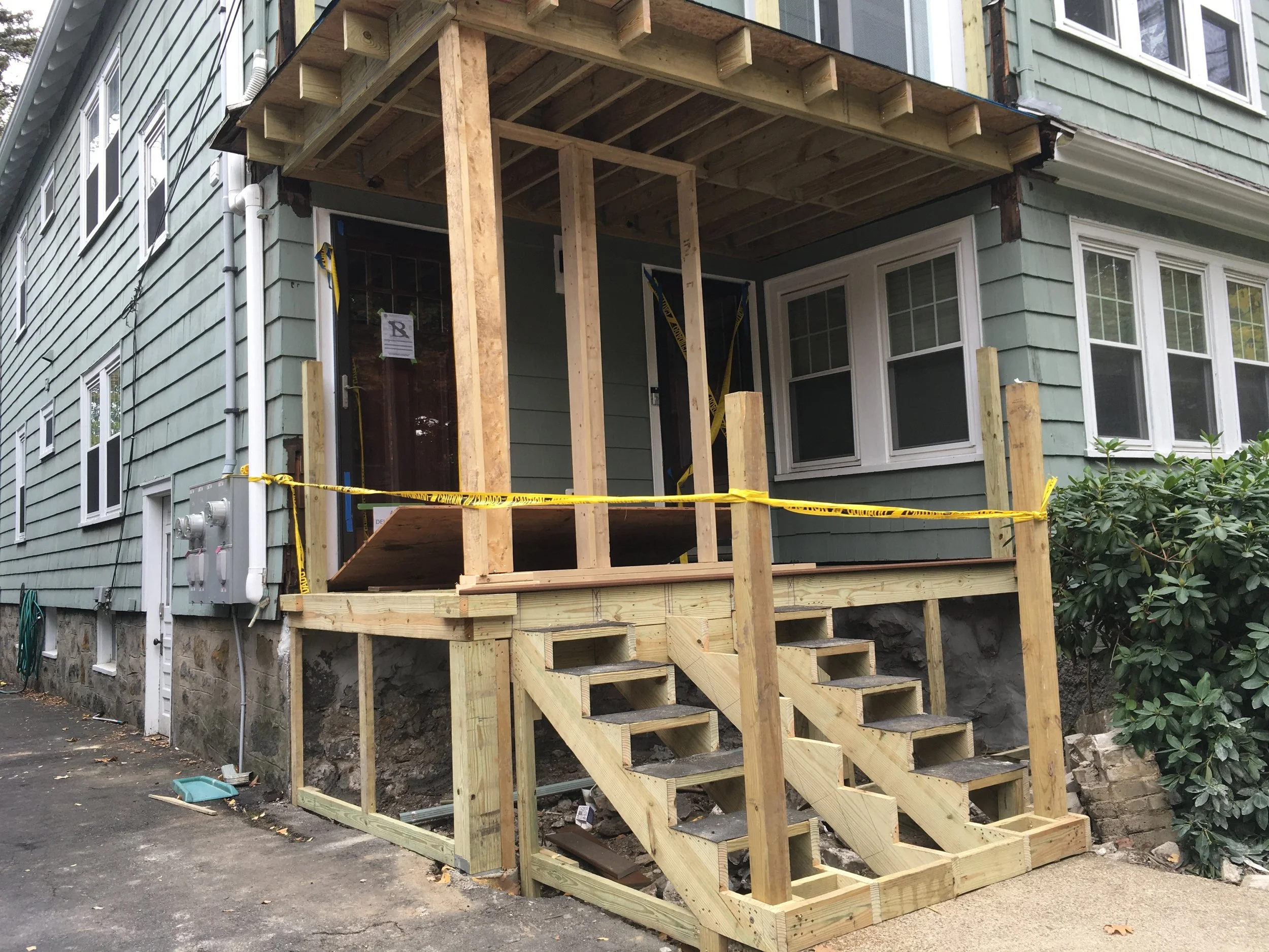 Construction of a new front porch on a house. The porch has a wooden frame, stairs, and a covered roof. Yellow caution tape surrounds the area.
