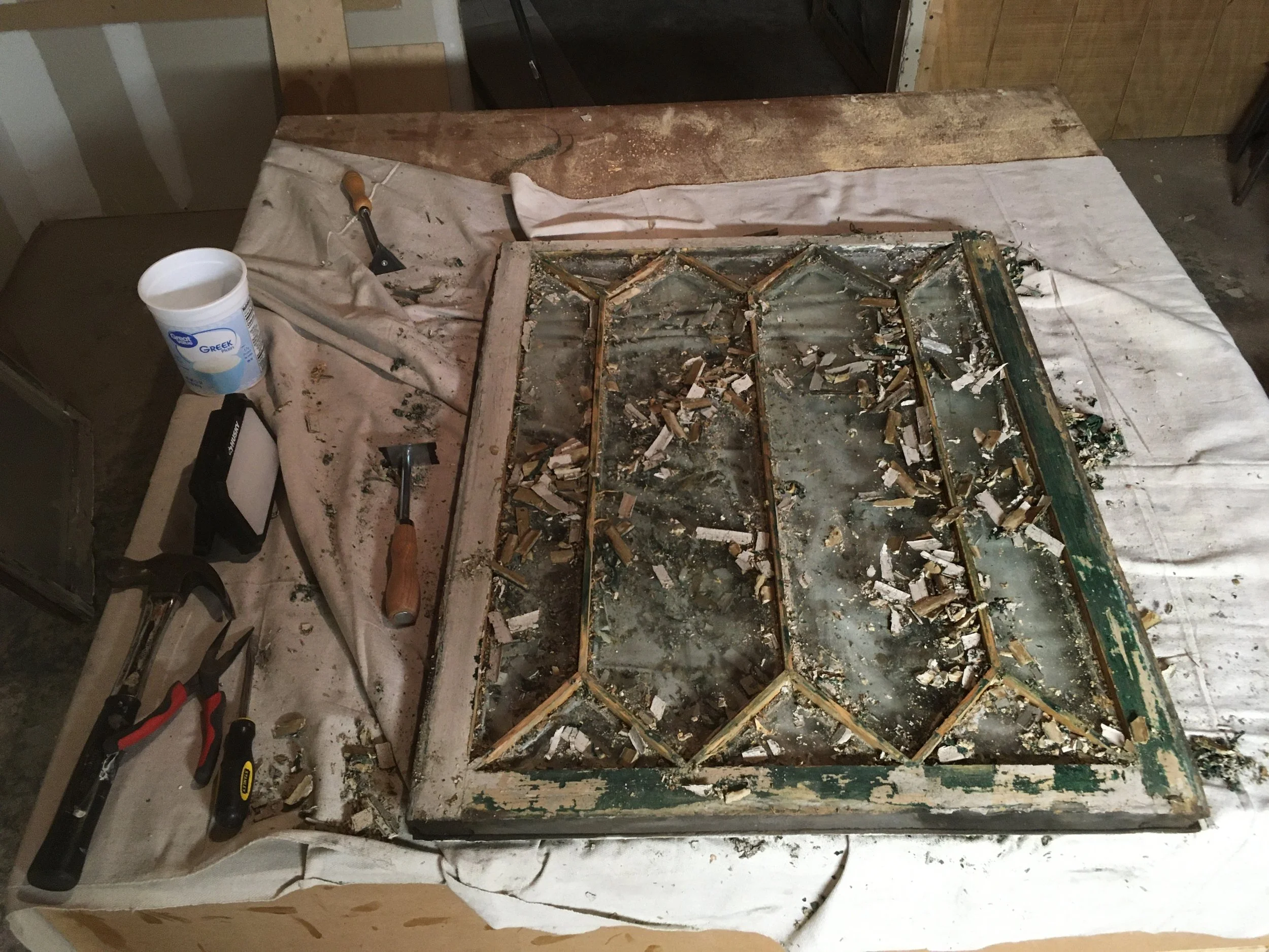 old, broken window with peeling paint and shattered glass on a worktable surrounded by tools and a container of cleaning solution.