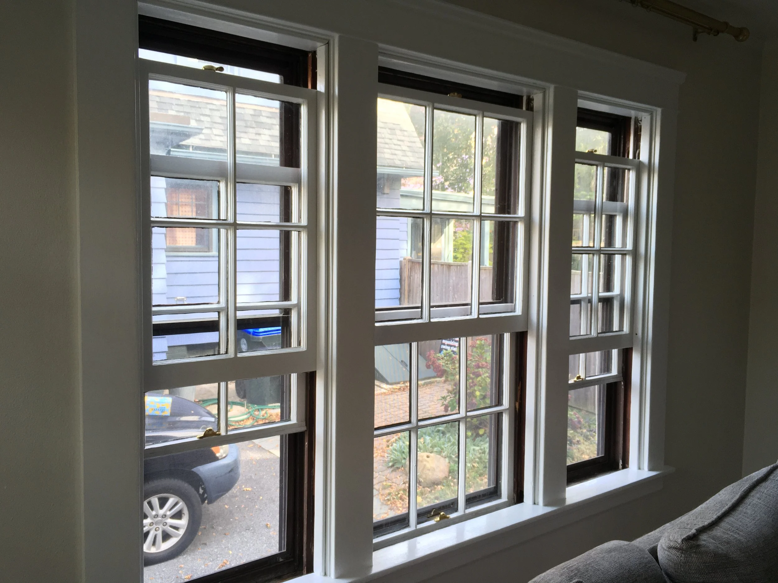 View of three double-hung windows with white muntins and dark wooden frames, showing a driveway with a parked car, a blue house, a wooden fence, and some trees outside.