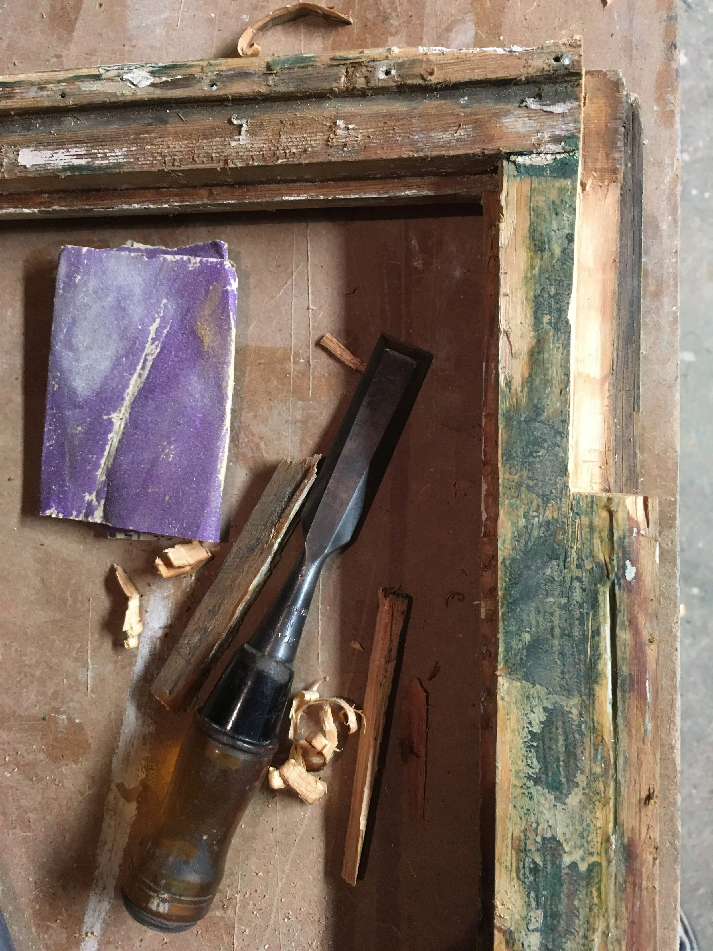 A woodworking workbench with a chisel, wood shavings, and a paint scraper, alongside pieces of wood and a sanding sponge.