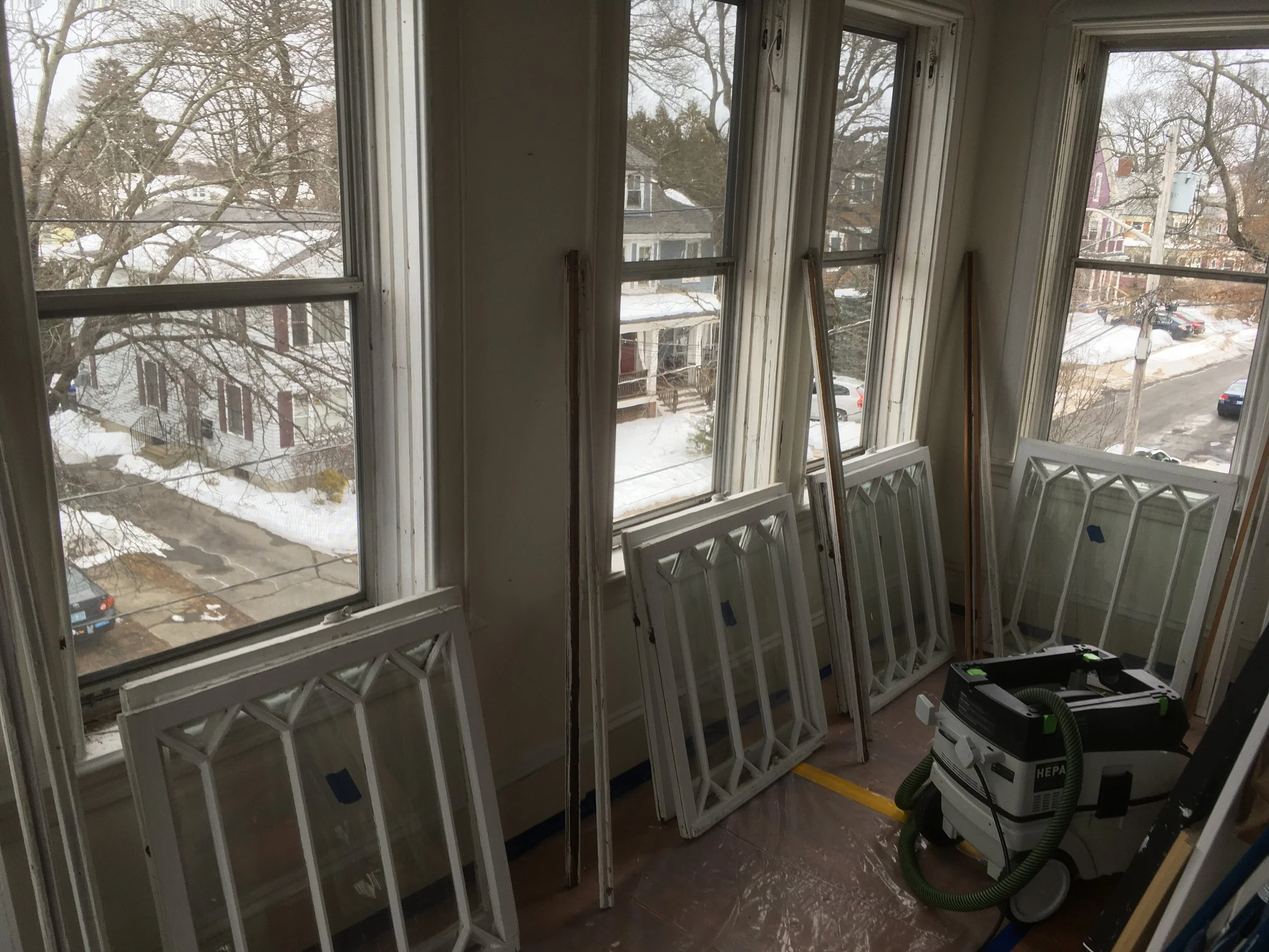A room with large windows showing a snowy residential street outside. Several window frames and glass panes are leaning against the walls, and a vacuum cleaner is on the floor, suggesting ongoing window replacement or renovation.