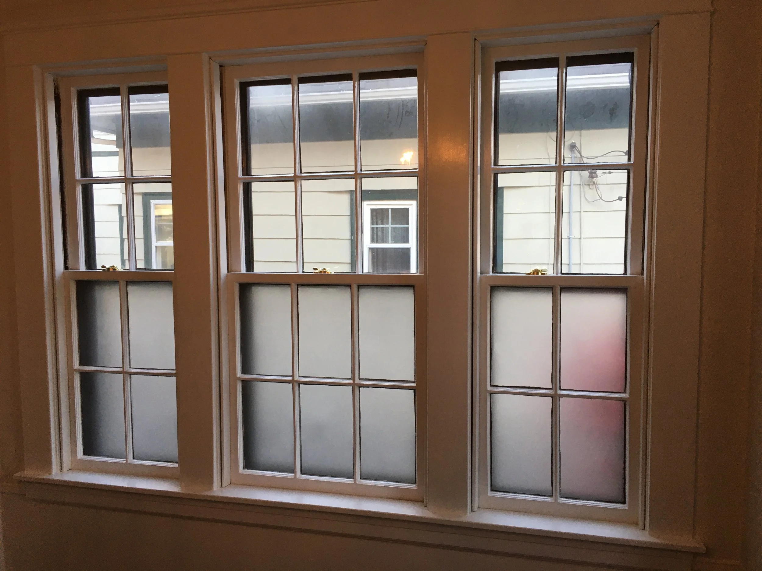 Three white-framed double-hung windows with grid patterns, frosted lower panes, and clear upper panes, set in a wall with a beige interior.