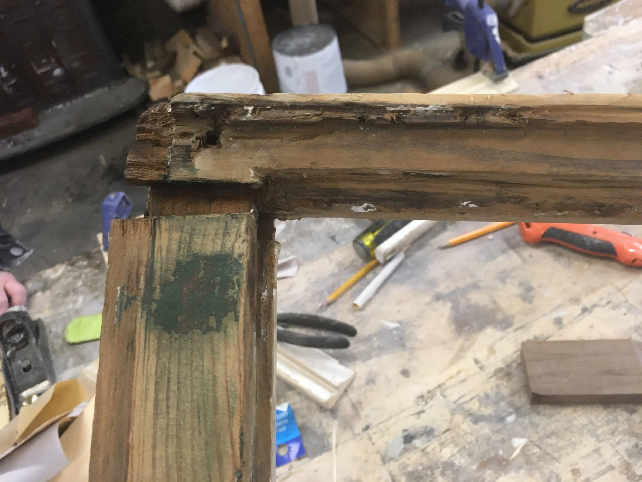 Close-up of a woodworking joint showing a damaged and worn wooden frame in a workshop. Tools and materials are scattered in the background.