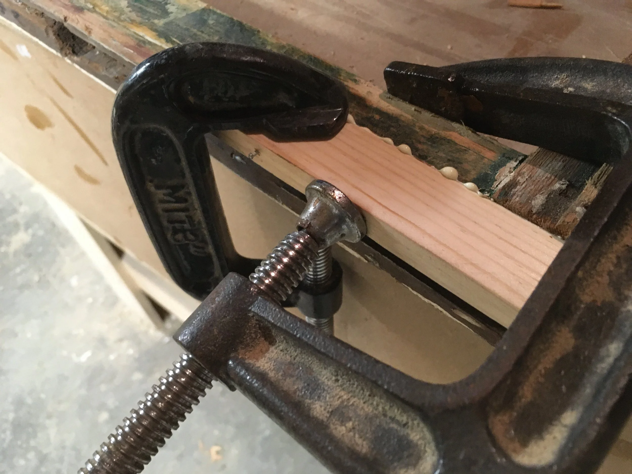 A close-up photo shows a woodworking clamp holding a small piece of light-colored wood to a workbench. The workbench is old and worn, with chipped paint and sawdust.