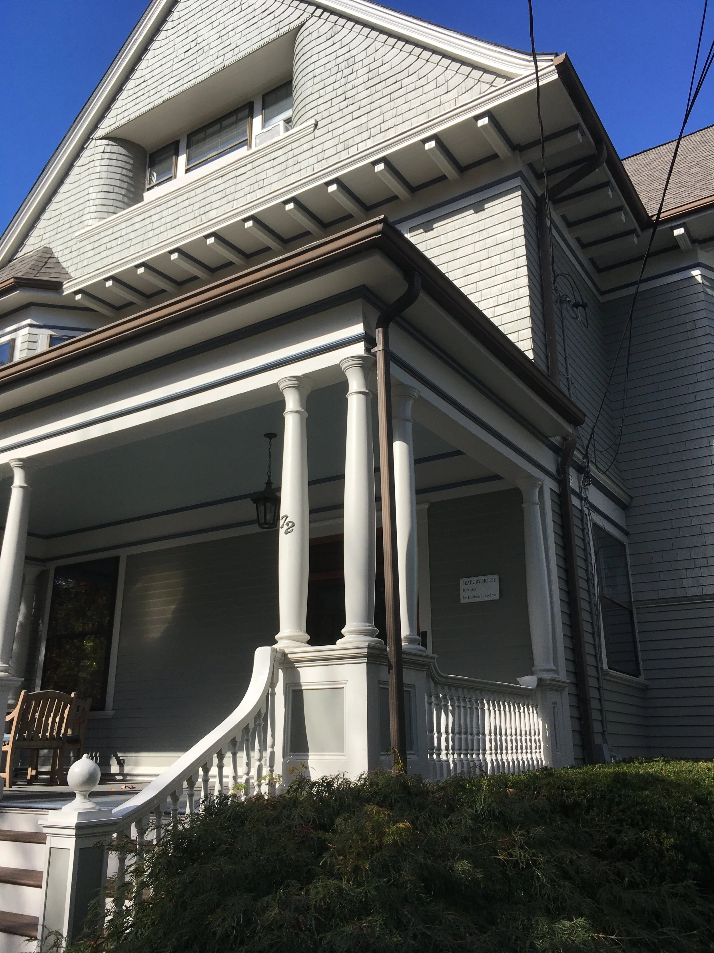 Front view of a large, Victorian-style house with a porch supported by tall columns, white wooden siding, and decorative trim, under a clear blue sky.