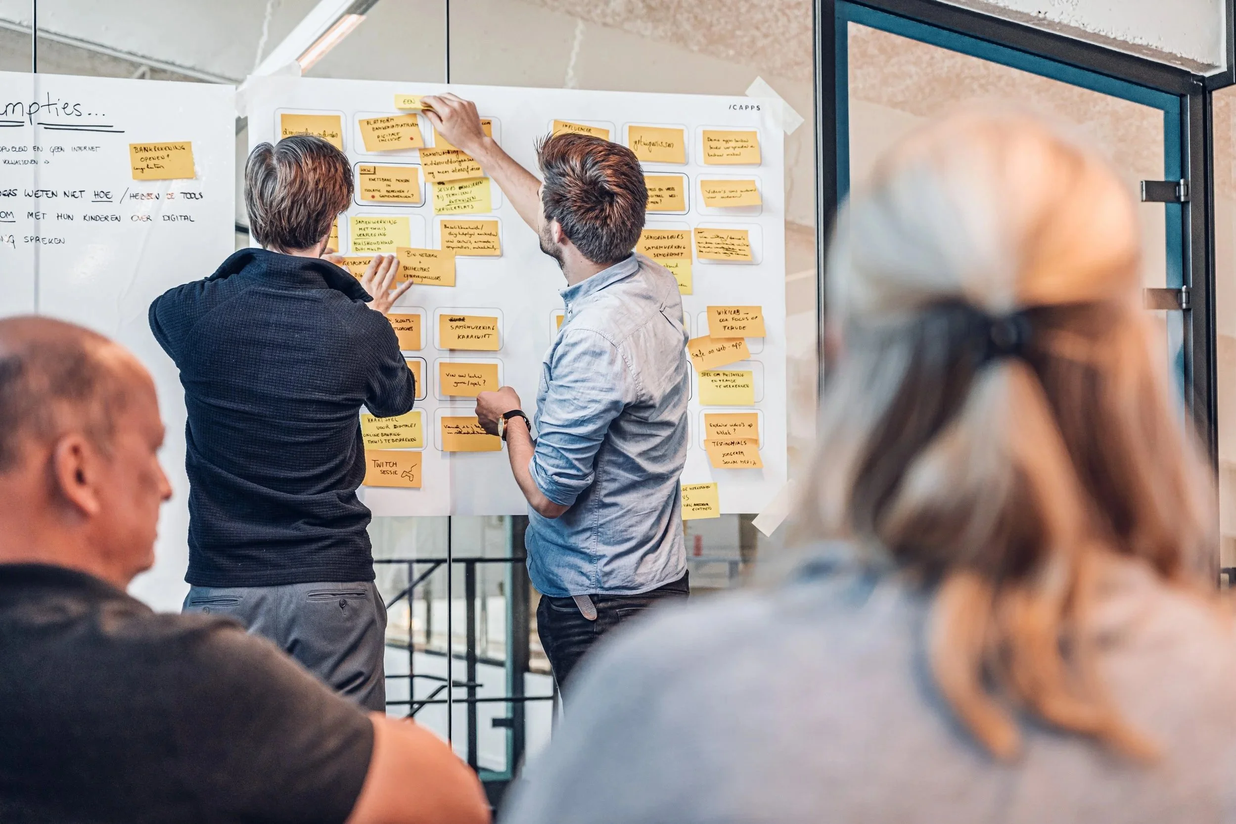 Two men are standing in front of a whiteboard covered with yellow sticky notes, collaborating on a project. Three other people are seated in front, watching the presentation in a modern office environment.