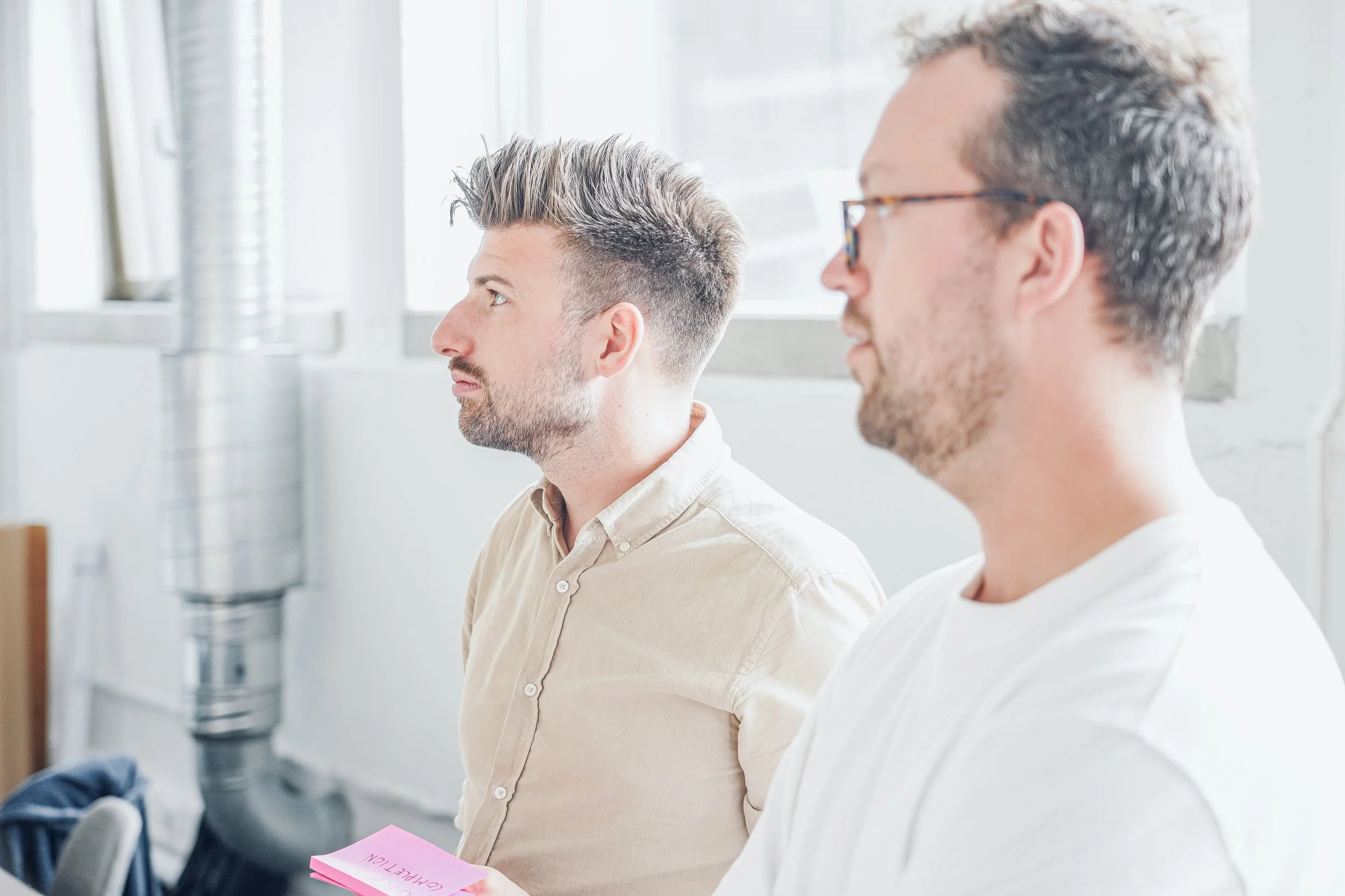 Two men sitting side by side in a meeting or class, one holding pink sticky notes.