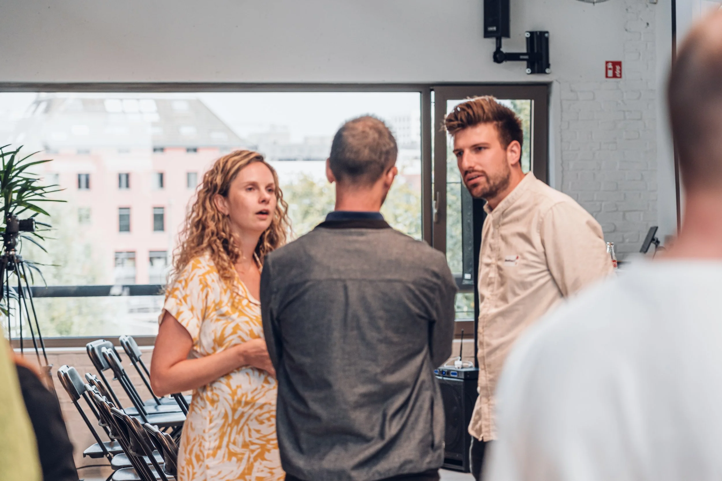 Three people are having a conversation indoors near large windows with a cityscape view in the background.