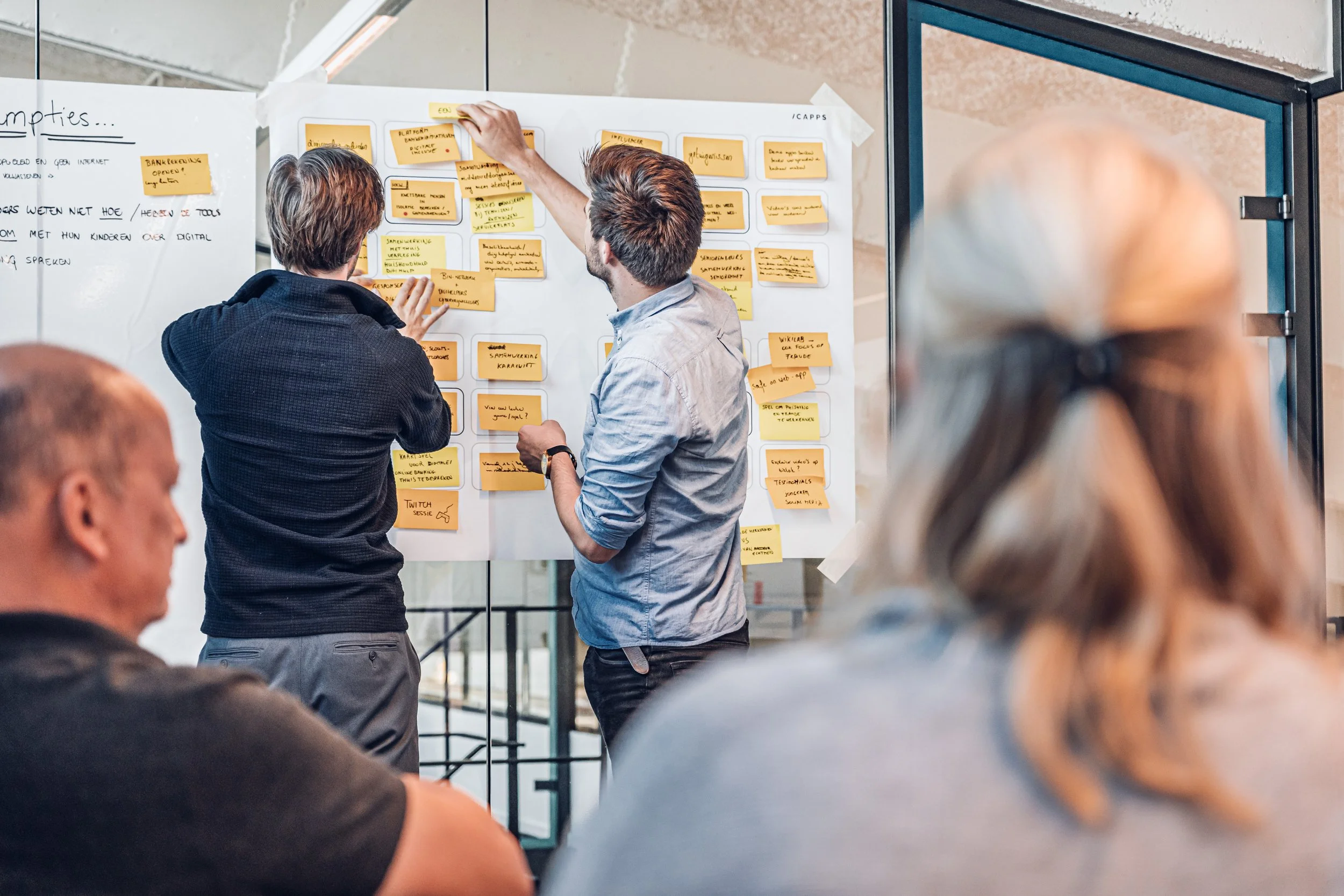 Two men presenting and discussing ideas on a whiteboard covered with yellow sticky notes in a meeting room, with others observing.