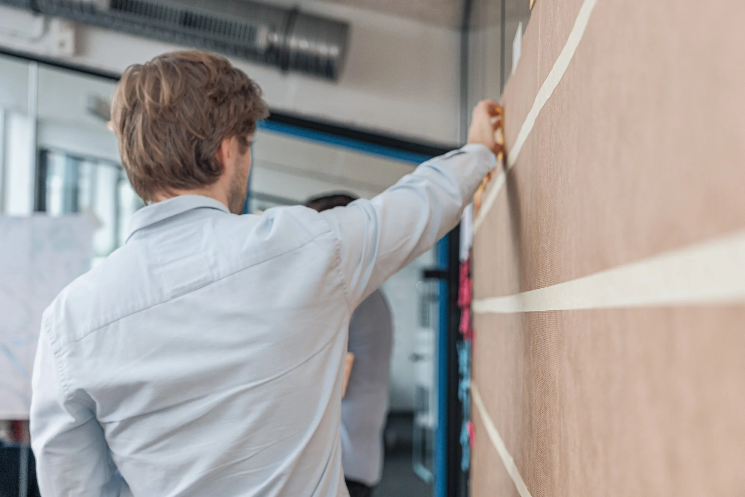 A man with brown hair in a white shirt is attaching papers or notes to a vertical brown board with horizontal white strips in an office setting.