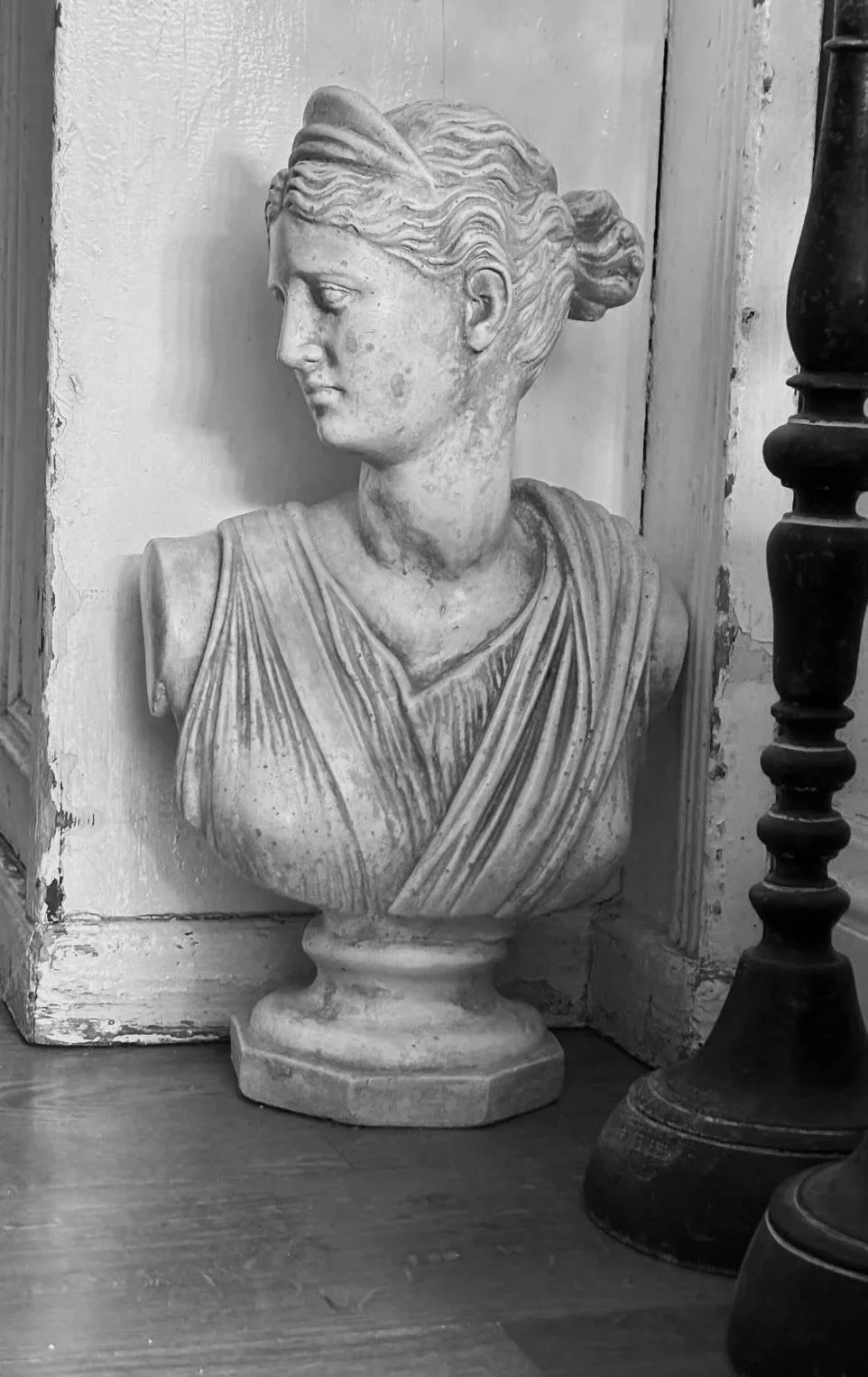 Black and white photograph of a classical marble bust of a woman with her hair in an updo, placed on a wooden surface next to a dark lamp.