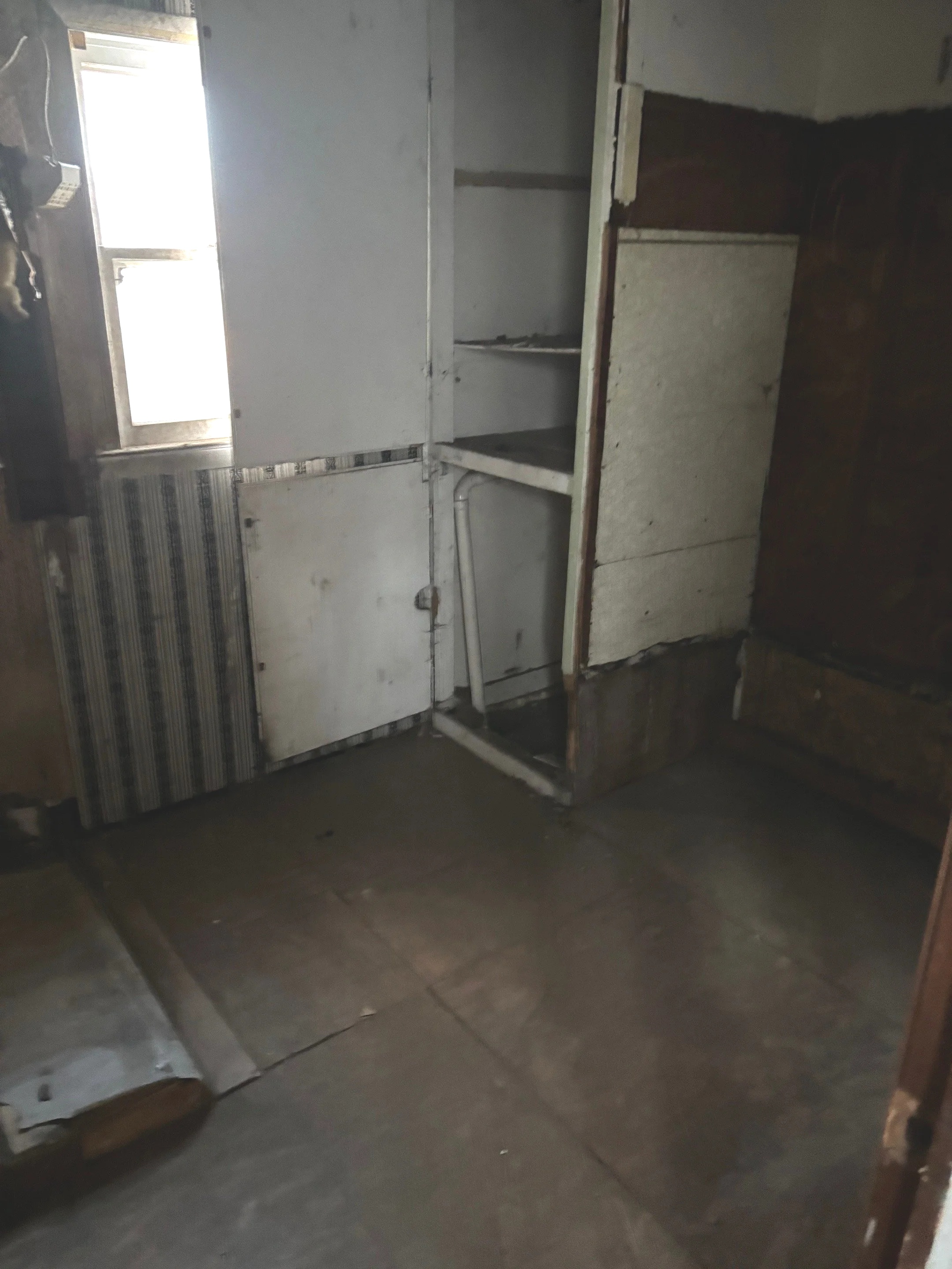 Empty and worn kitchen with exposed drywall and flooring, small window, and open closet or cabinet space.