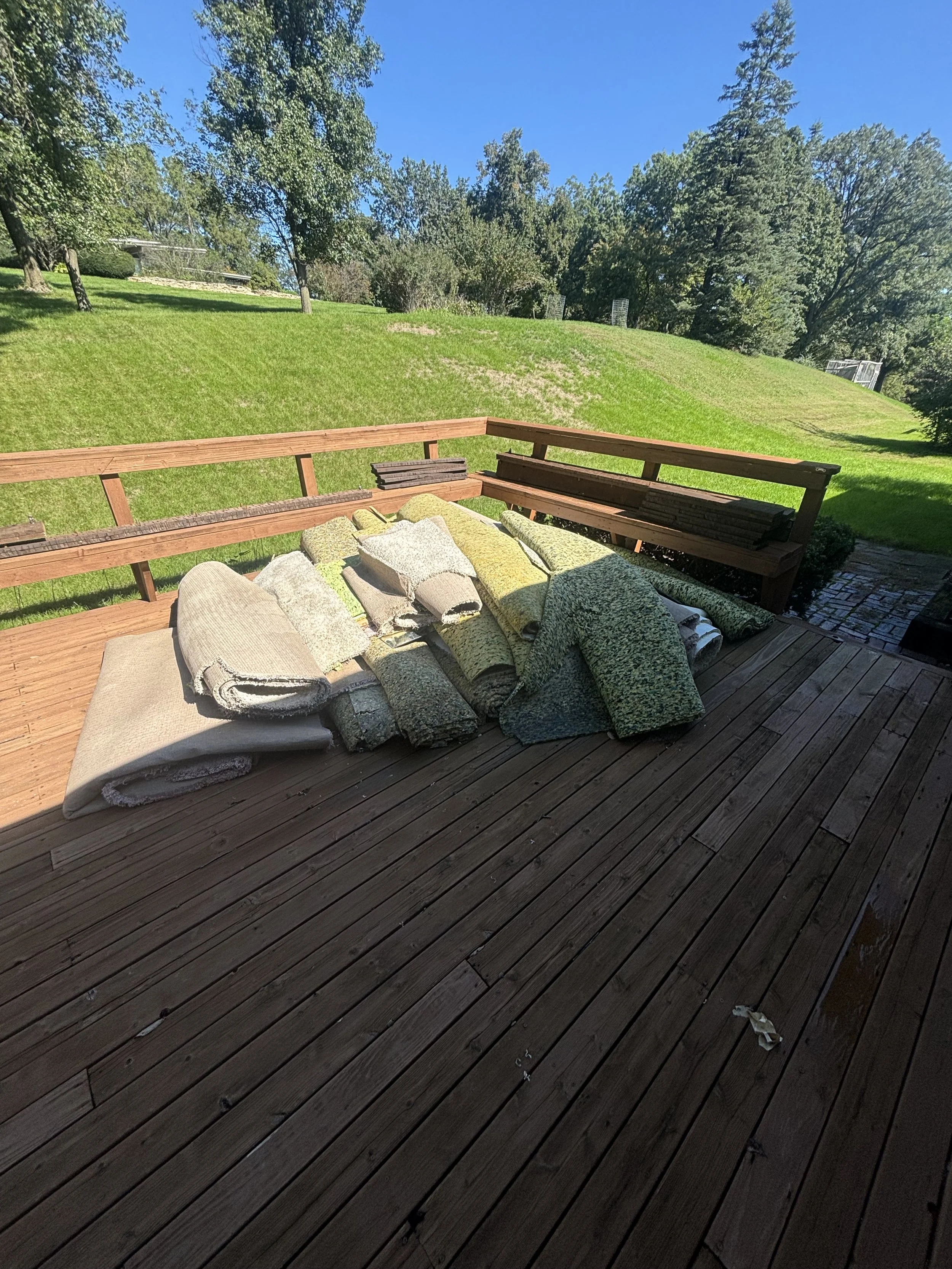 A wooden deck with a pile of rolled-up carpets or rugs in various colors and patterns. In the background, a grassy hill with trees and a clear blue sky.
