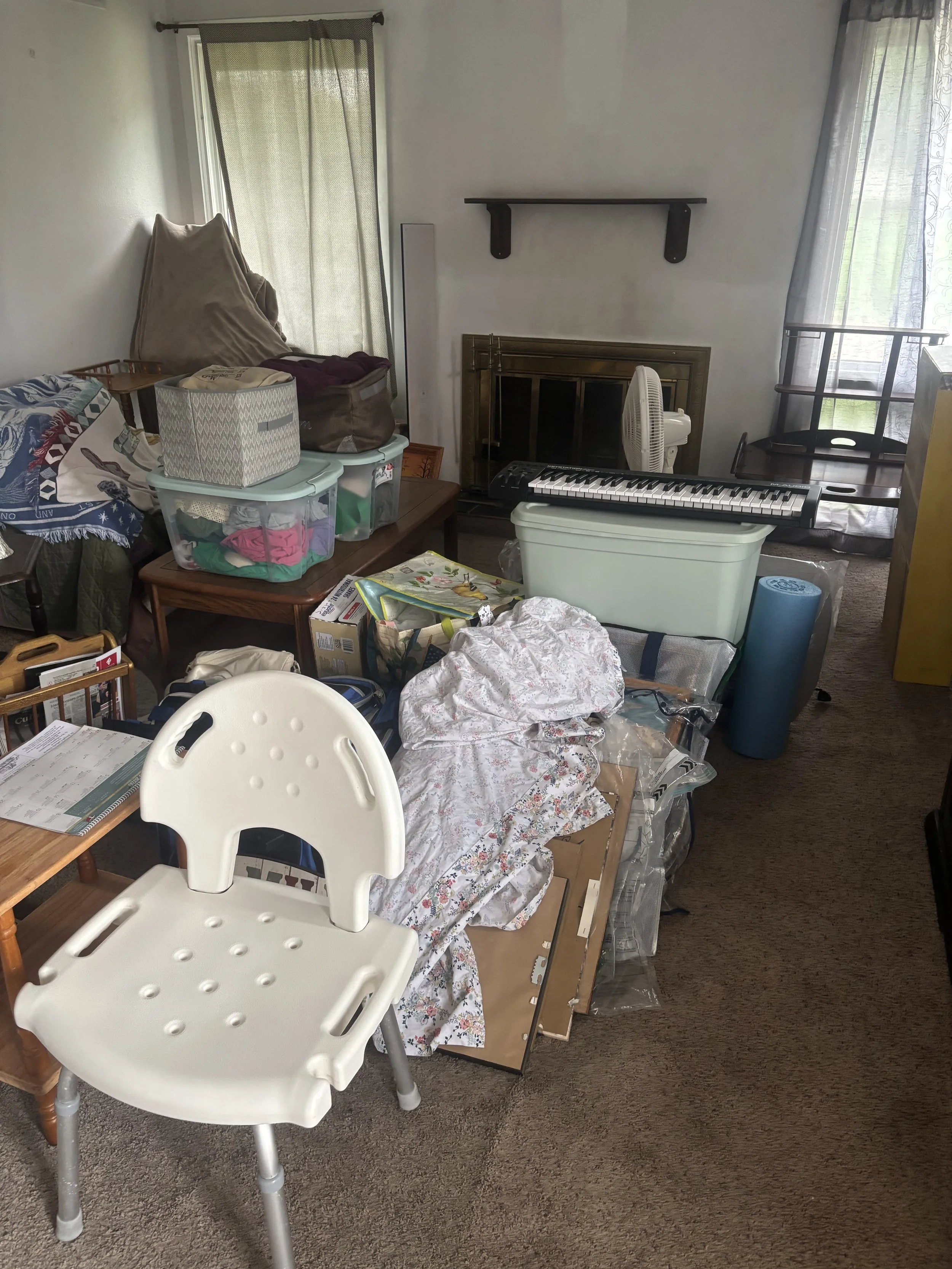 A cluttered living room with plastic storage bins, a white high chair, a piano keyboard, a fan, a fireplace, and various furniture and items in disarray.